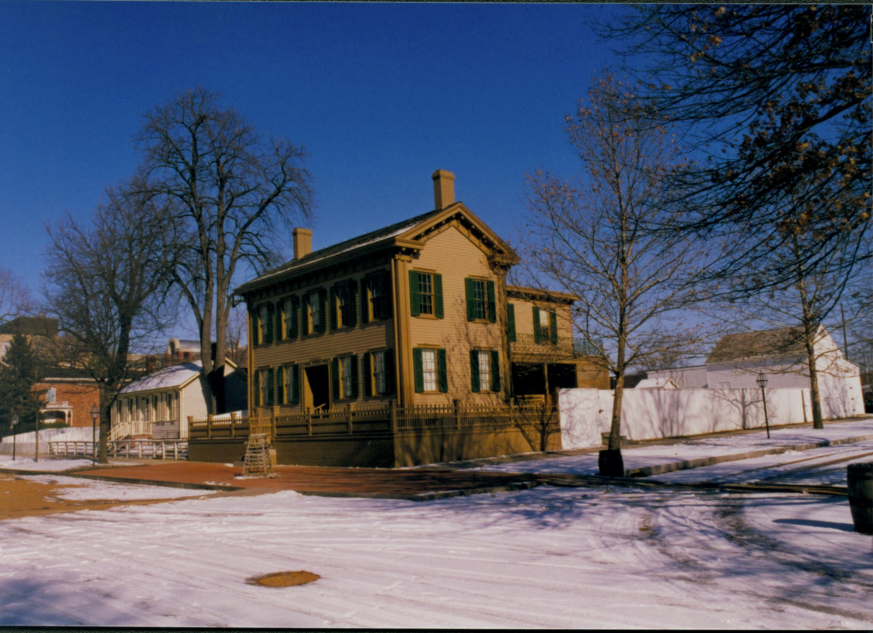 Lincoln neighborhood in winter with some snow.  Lincoln Home in center with cleared brick plaza in front and elm tree in cage on plaza.  Corneau House on left, Conference Center in brick on far left.  Lincoln Barn on right behind white board fence. Trash barrels on corners of 8th and Jackson Street intersection. Manhole cover cleared of snow in center of intersection Looking Northeast from 8th and Jackson Street intersection snow, Lincoln Home, Corneau, Conference Center, Lincoln Barn, 8th Street, Jackson Street