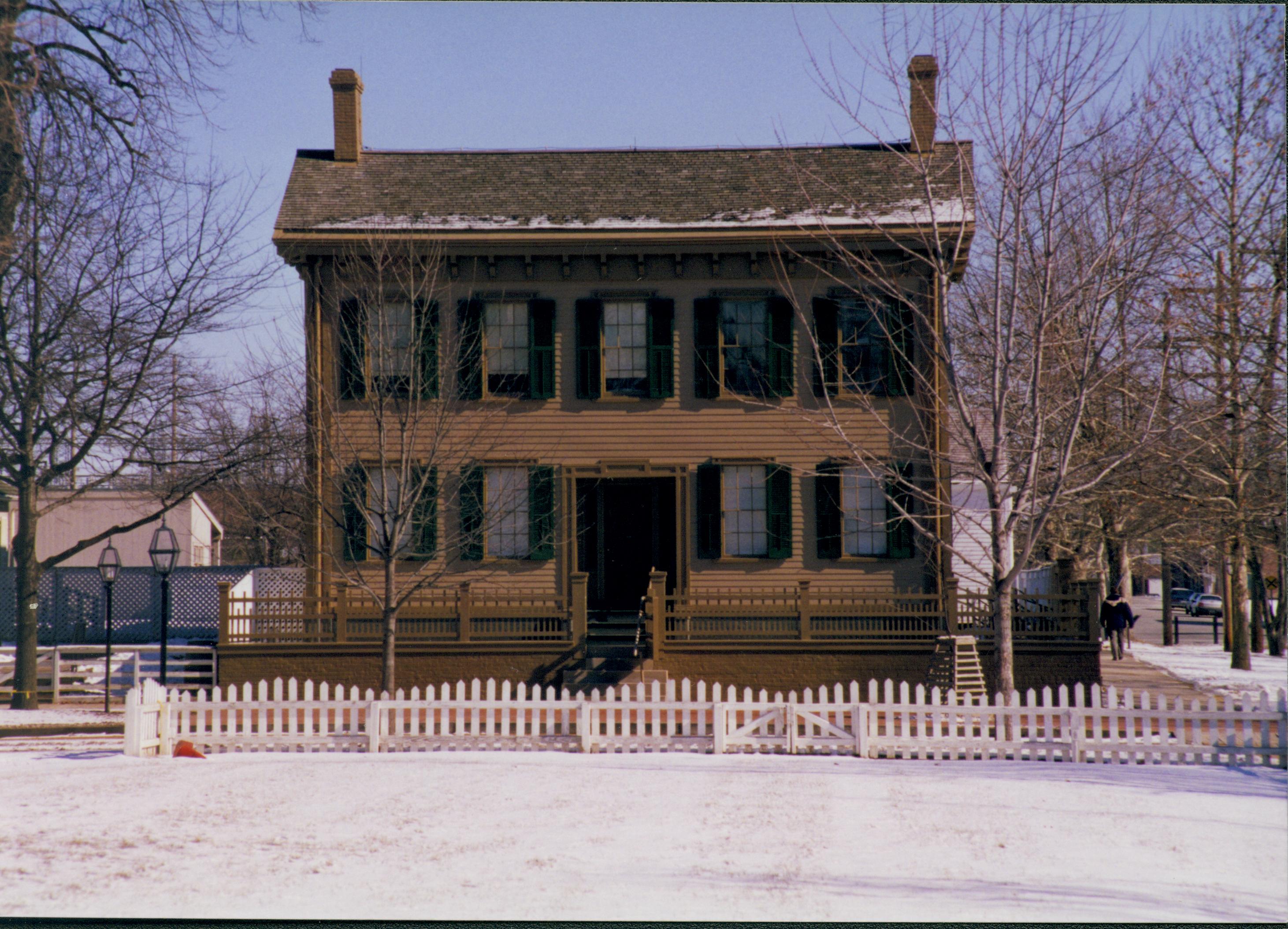 Lincoln Home in snow, from Burch lot, elm tree in cage on right. B-2 in background left, corner of Corneau House on far left. Fence from Burch Lot in foreground. Cleared boardwalk visible on right with visitors walking away Looking East from Burch Lot, Block 7, Lot 9 snow, Lincoln Home, Burch Lot, B-2, Corneau, elm tree