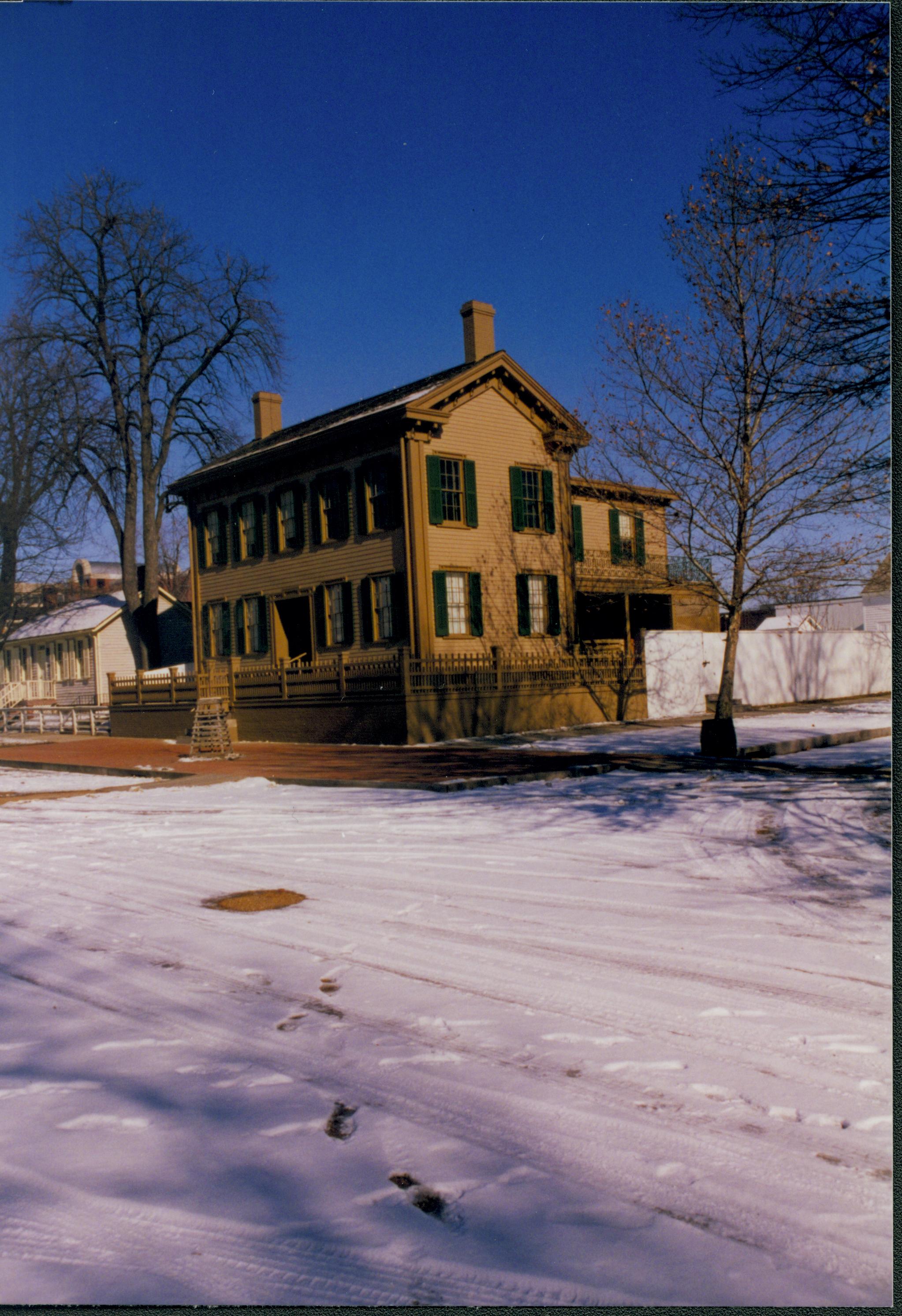 Lincoln Home in snow, cleared brick plaza in front with elm tree in cage on plaze. Corneau House on left with Sangamon County Courthouse in background over Corneau roof. Footprints, maintenance vehicle tracks, and manhole cover without snow visible in intersection of 8th and Jackson Streets. Cleared boardwalk along Jackson Street. Trash barrel on corner near Lincoln Home. Looking Northeast from 8th and Jackson Street intersection snow, Lincoln Home, brick plaza, elm tree, 8th Street, Jackson Street, Corneau, boardwalks