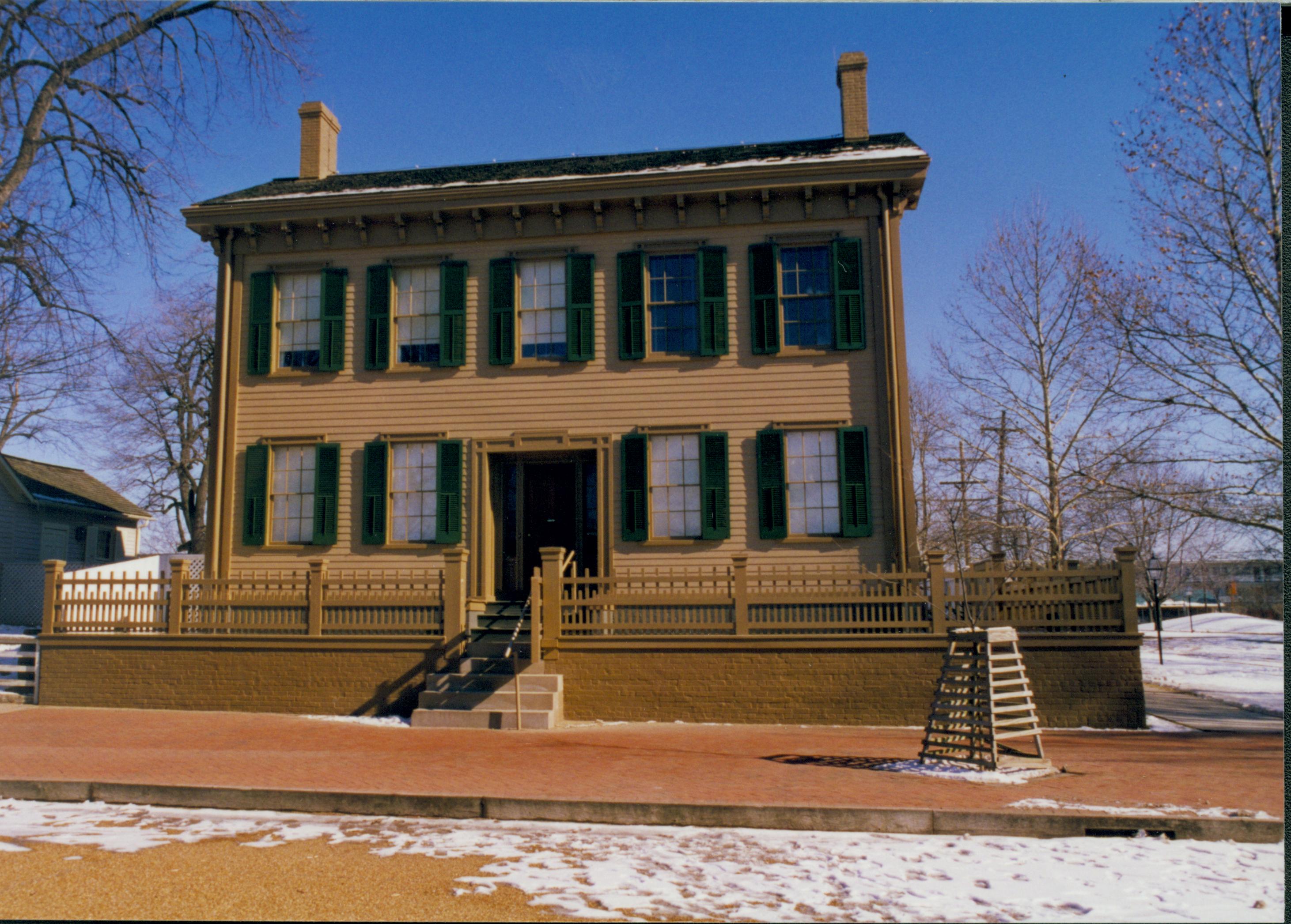 Lincoln Home in winter with some snow, cleared brick plaza in front, elm tree in cage on right near cleared boardwalk on far right. Corneau House on left. Looking East from 8th Street snow, Lincoln Home, brick plaza, elm tree, 8th Street, Corneau