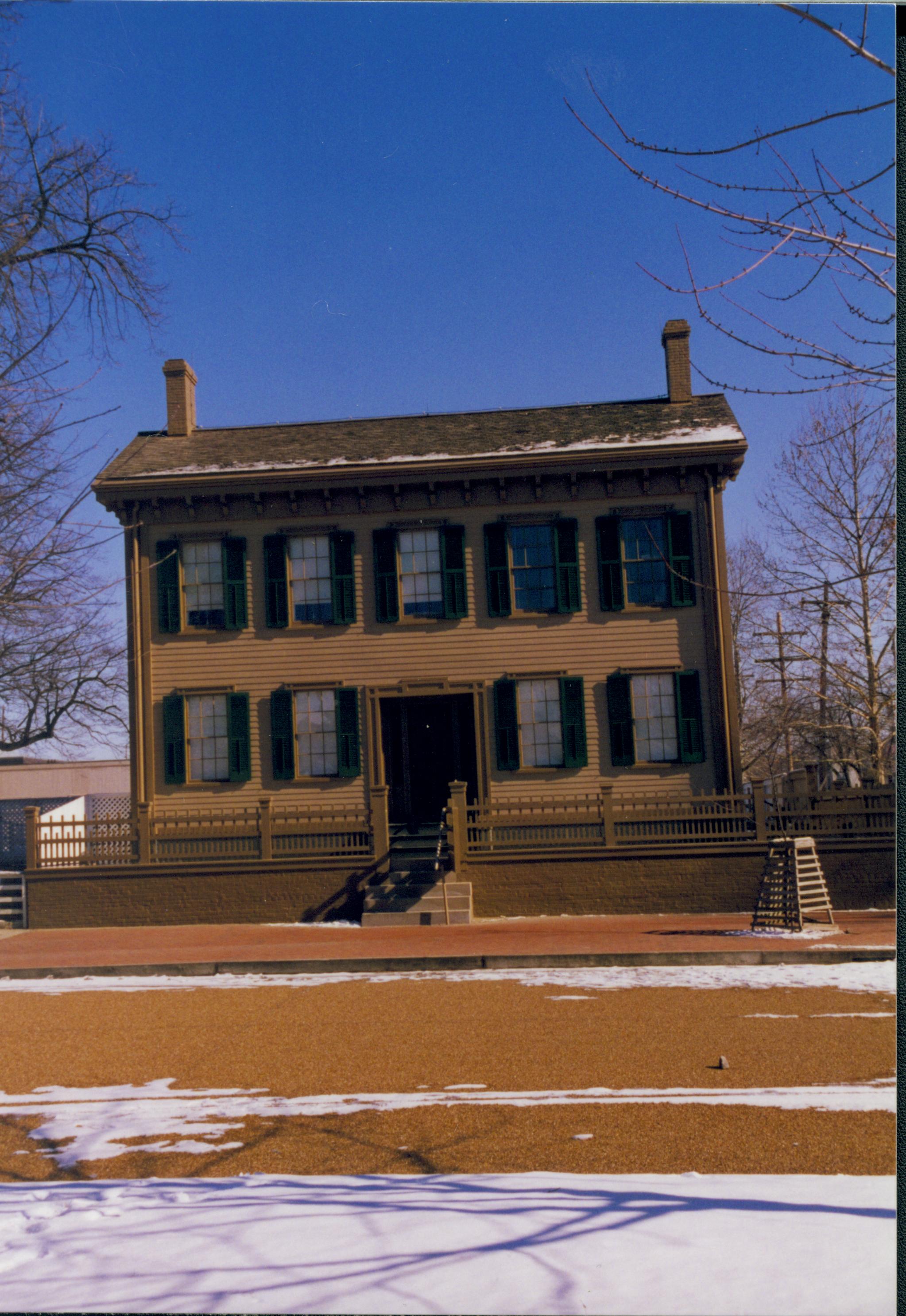 Lincoln Home in winter with some snow, cleared brick plaza in front with elm tree in cage on right. B-2 in background on left. Looking East from west side of 8th Street snow, Lincoln Home, brick plaza, elm tree, 8th Street, B-2