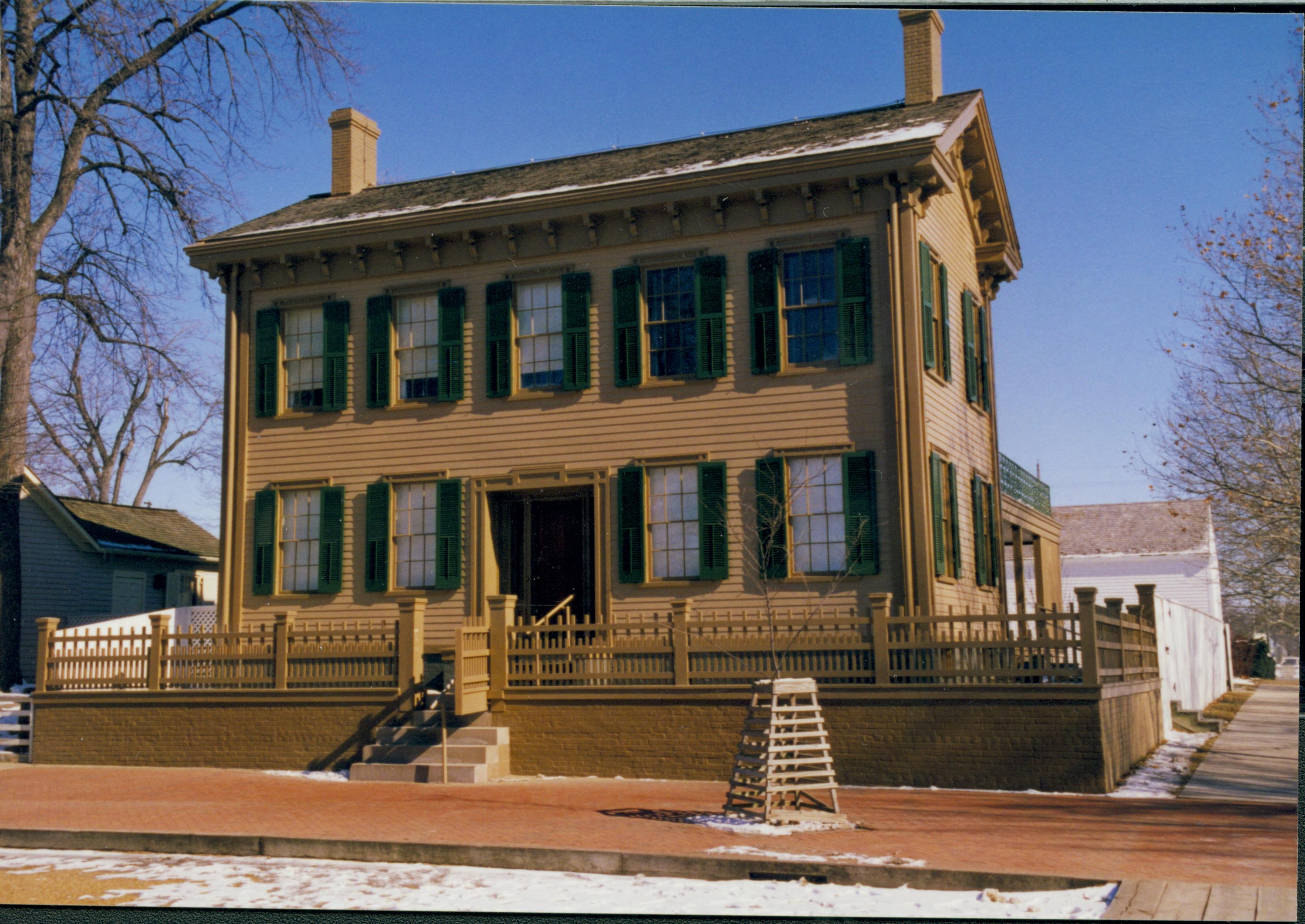 Lincoln Home in winter with some snow, cleared brick plaza in front, elm tree in cage in foreground right. Corneau House on left, Lincoln Barn in background right.  Cleared boardwalk on far right. Looking East/Northeast from 8th and Jackson Street intersection snow, Lincoln Home, brick plaza, elm tree, Corneau, Lincoln Barn, boardwalk, 8th Street, Jackson Street