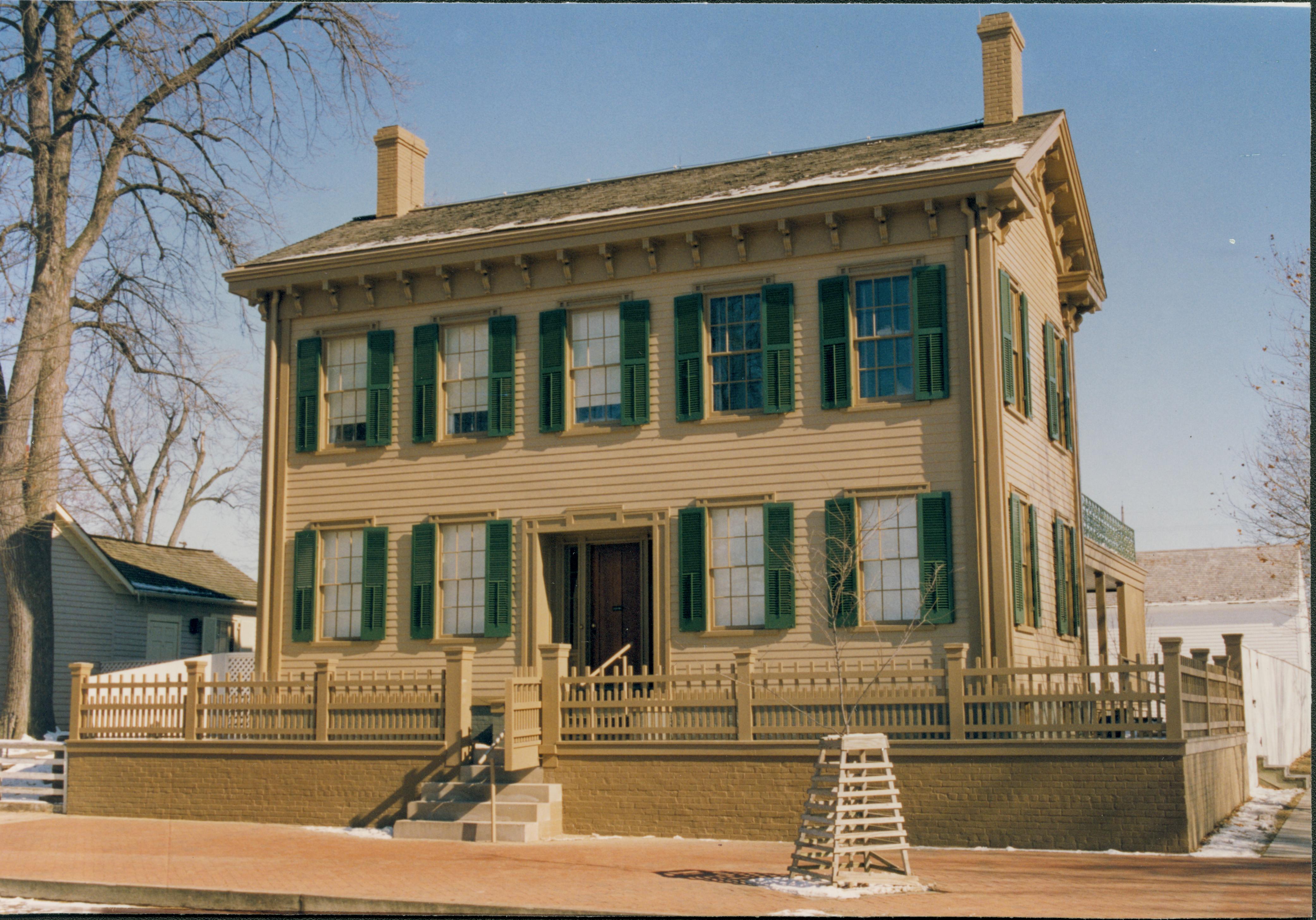 Lincoln Home in winter with some snow, cleared brick plaza, elm tree in cage on right.  Corneau House is on left. Lincoln Barn is in the background on the far right. Looking East/Northeast from the 8th and Jackson Streets intersection snow, Lincoln Home, Corneau, Lincoln Barn, 8th Street, brick plaza, elm tree