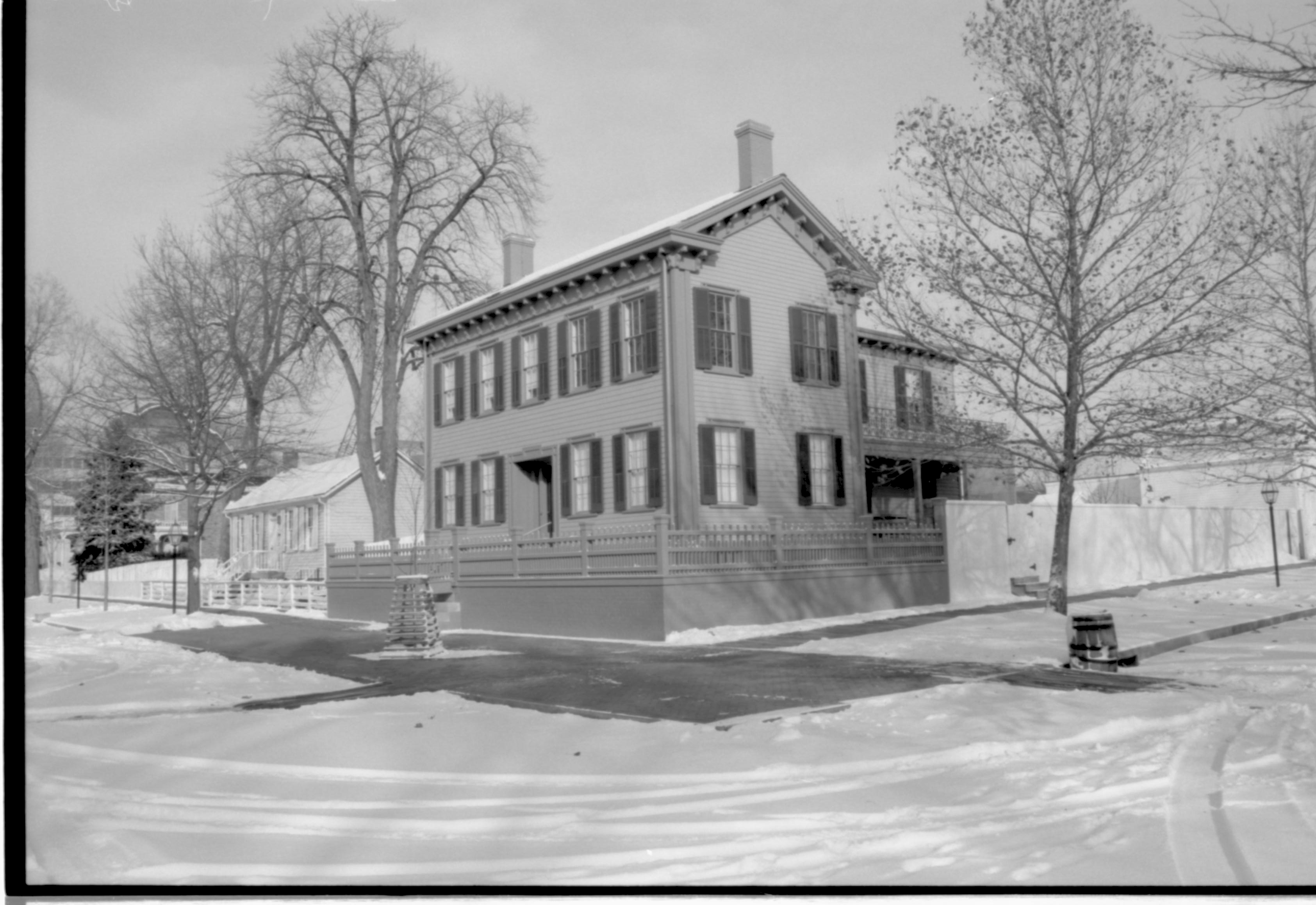 Lincoln Home in snow, cleared brick plaza in front, elm tree in cage on plaza.  Corneau House on left, Conference Center in brick on far left. Lincoln Barn and Woodshed on right behind fence. Trash barrel in gutter along Jackson Street. Maintenance vehicle tracks visible in streets Looking Northeast from 8th and Jackson Street intersection snow, Lincoln Home, Corneau, Lincoln Barn, Lincoln Woodshed, Conference Center, brick plaza, elm tree, fence