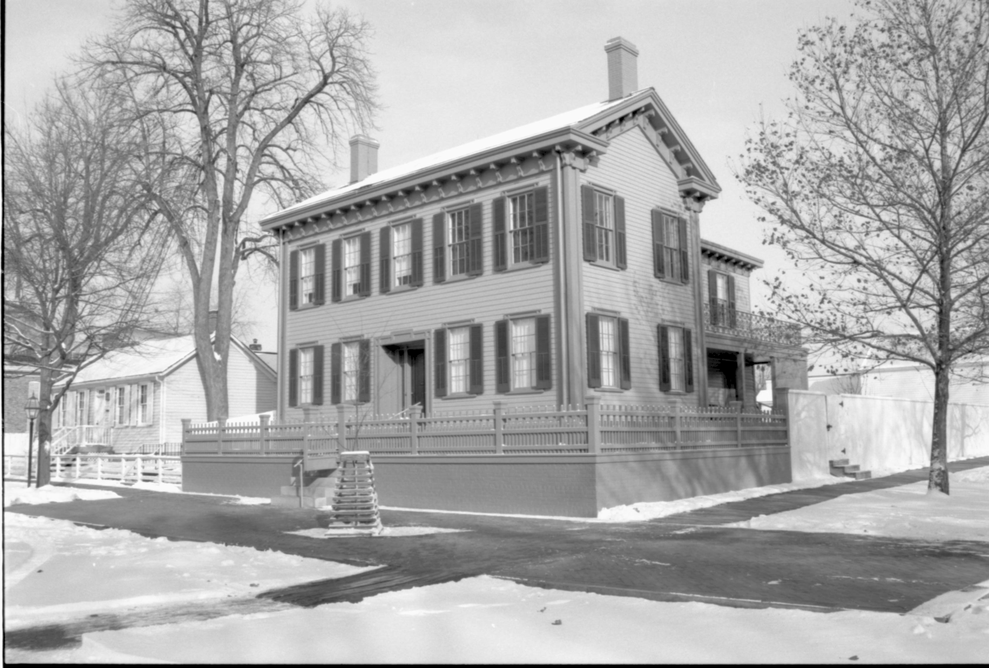 Lincoln Home in the snow, cleared brick plaza in front, elm tree in cage on plaza.  Corneau House on left, Conference Center in brick on far left.  Cleared boardwalk along fence on right, Lincoln Barn on far right behind tree. Looking Northeast from 8th and Jackson Street intersection snow, Lincoln Home, Corneau, Conference Center, Lincoln Barn, 8th Street, Jackson Street, boardwalk, brick plaza, elm tree