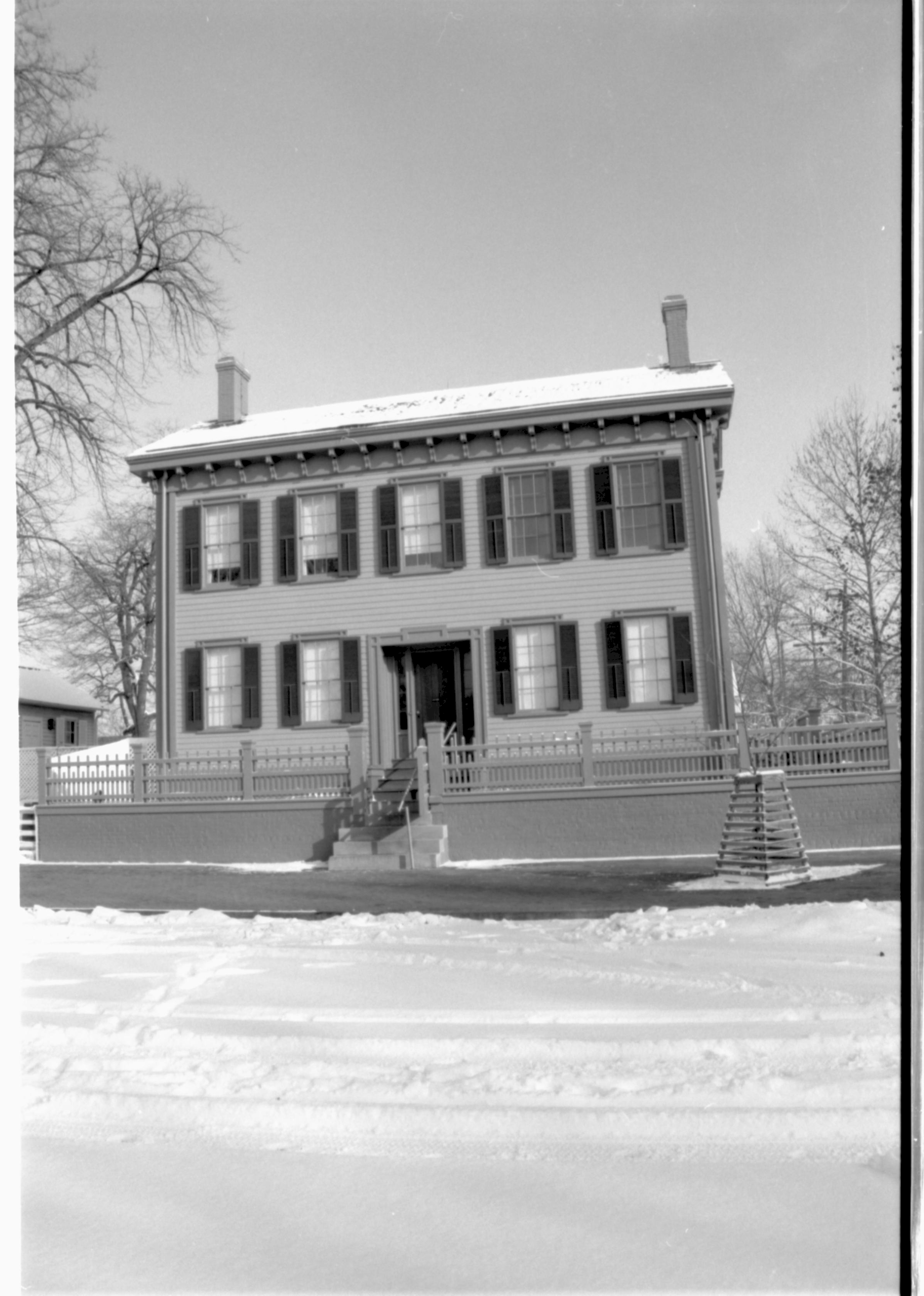 Lincoln Home in snow, cleared brick plaza in front, elm tree in cage on right. Corneau House on far left. Maintenance vehicle tracks in snow in foreground. Looking East from 8th Street snow, Lincoln Home, Corneau, brick plaza, elm tree, 8th Street