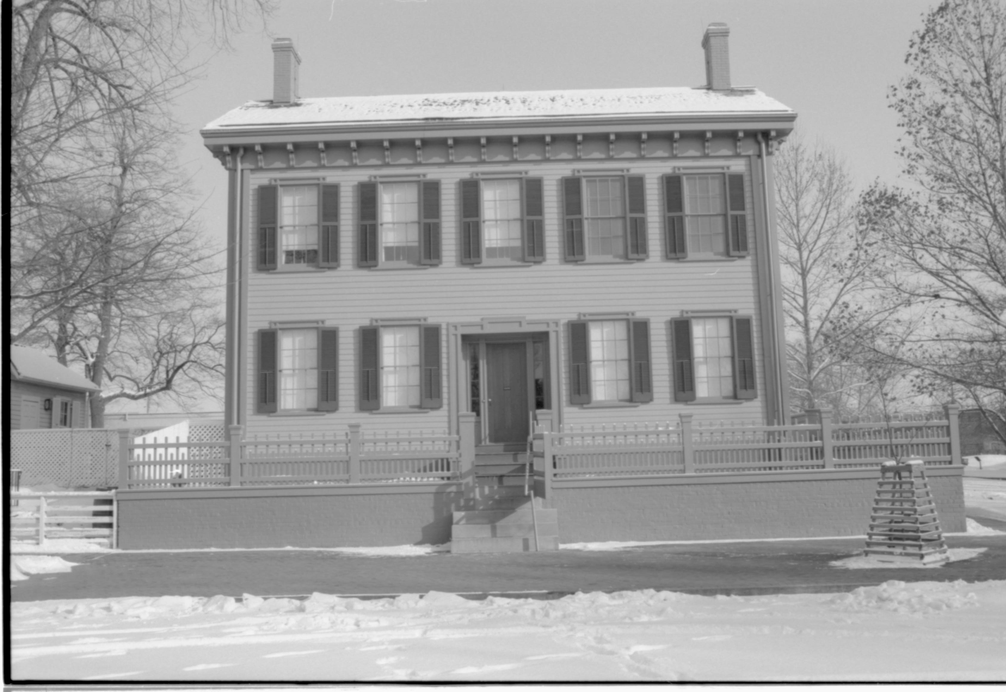 Lincoln Home in snow, cleared brick plaza in front, elm tree in cage on right.  Corneau House on far left. Maintenance vehicle tracks and footprints in snow in foreground. Looking East from 8th Street snow, Lincoln Home, Corneau, brick plaza, elm tree, 8th Street