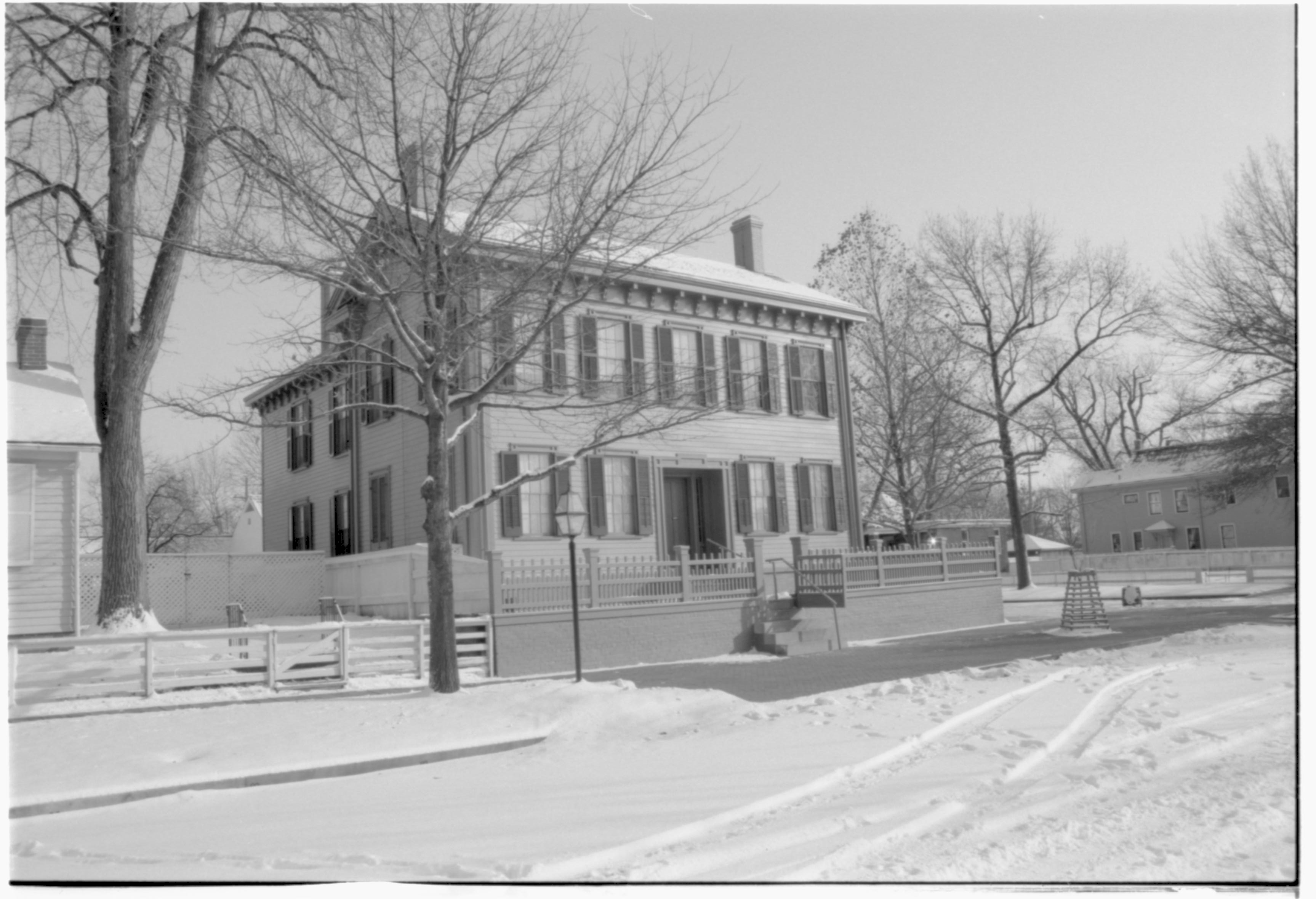 Lincoln Home in the snow, cleared brick plaza in front, elm tree in cage on right.  Arnold House in background right, Cook House on far right.  Corneau House on left, Lincoln Barn in background left behind Home. Maintenance vehicle tracks and footprints in snow in foreground Looking East/Southeast from boardwalk on west side of 8th Street snow, Lincoln Home, Corneau, Arnold, Cook, brick plaza, elm tree, Lincoln Barn, 8th Street