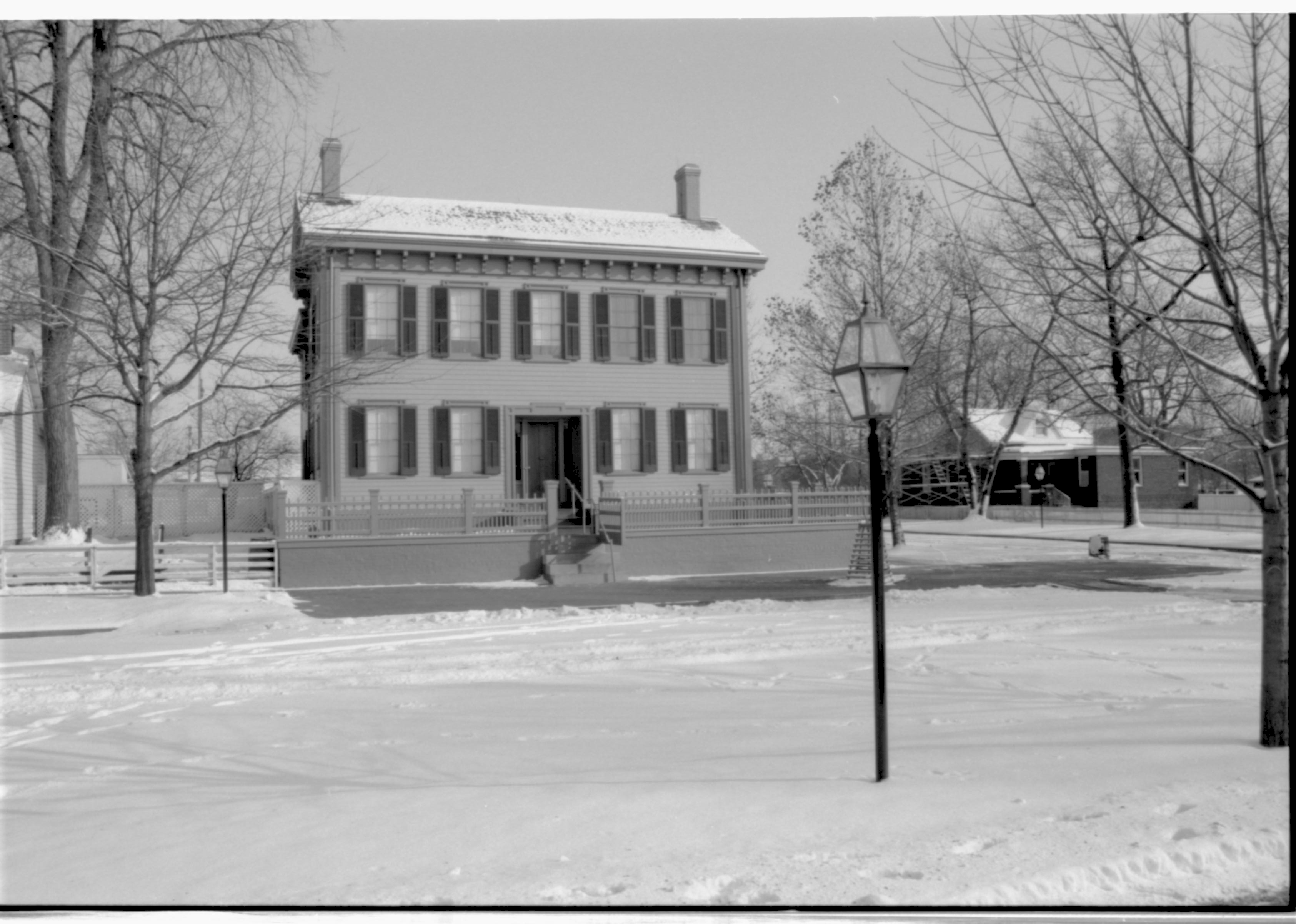 Lincoln Home in snow, cleared brick plaza in front, elm tree in cage on right behind street light.  Arnold House in background on right. Corneau House on far left. Looking East/Southeast from boardwalk west side of 8th Street snow, Lincoln Home, Corneau, Arnold, brick plaza, elm tree, 8th Street