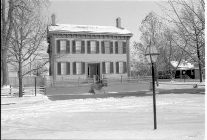 Lincoln Home in snow (blurry), cleared brick plaza in front, elm tree in cage behind street light. Arnold House in background right. Maintenance vehicle tracks and footprints in snow in foreground. Looking East/Southeast from boardwalk on west side of 8th Street snow, Lincoln Home, Arnold, brick plaza, elm tree, 8th Street