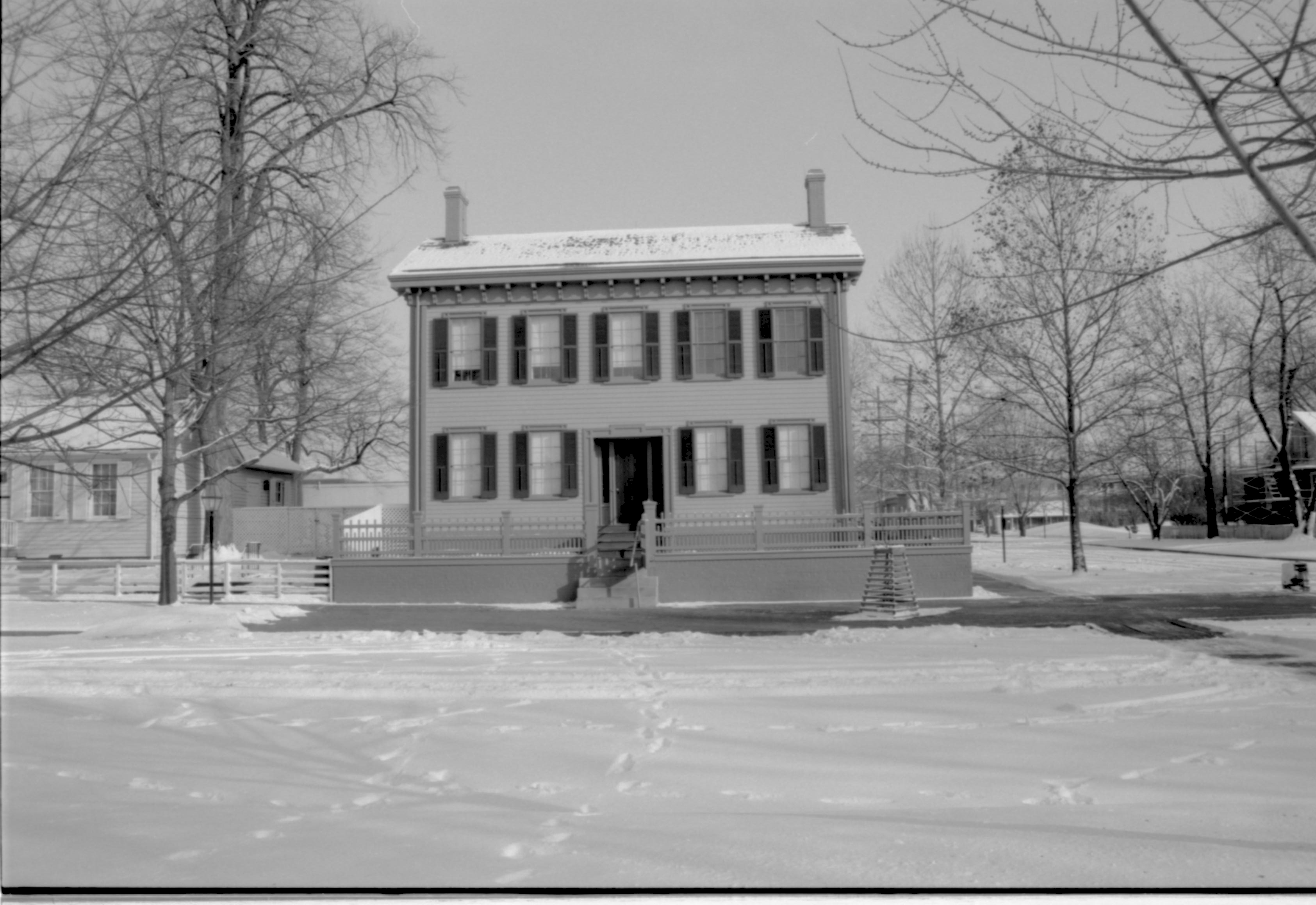 Lincoln Home in snow, cleared brick plaza in front, elm tree in cage on right. Corneau House on left. Arnold House with scaffolding on far right.  Former Travel Lodge Motel in background right. Looking East from Burch lot, Block 7, Lot 9 snow, Lincoln Home, Corneau, Arnold, Travel Lodge Motel, brick plaza, elm tree, Burch Lot