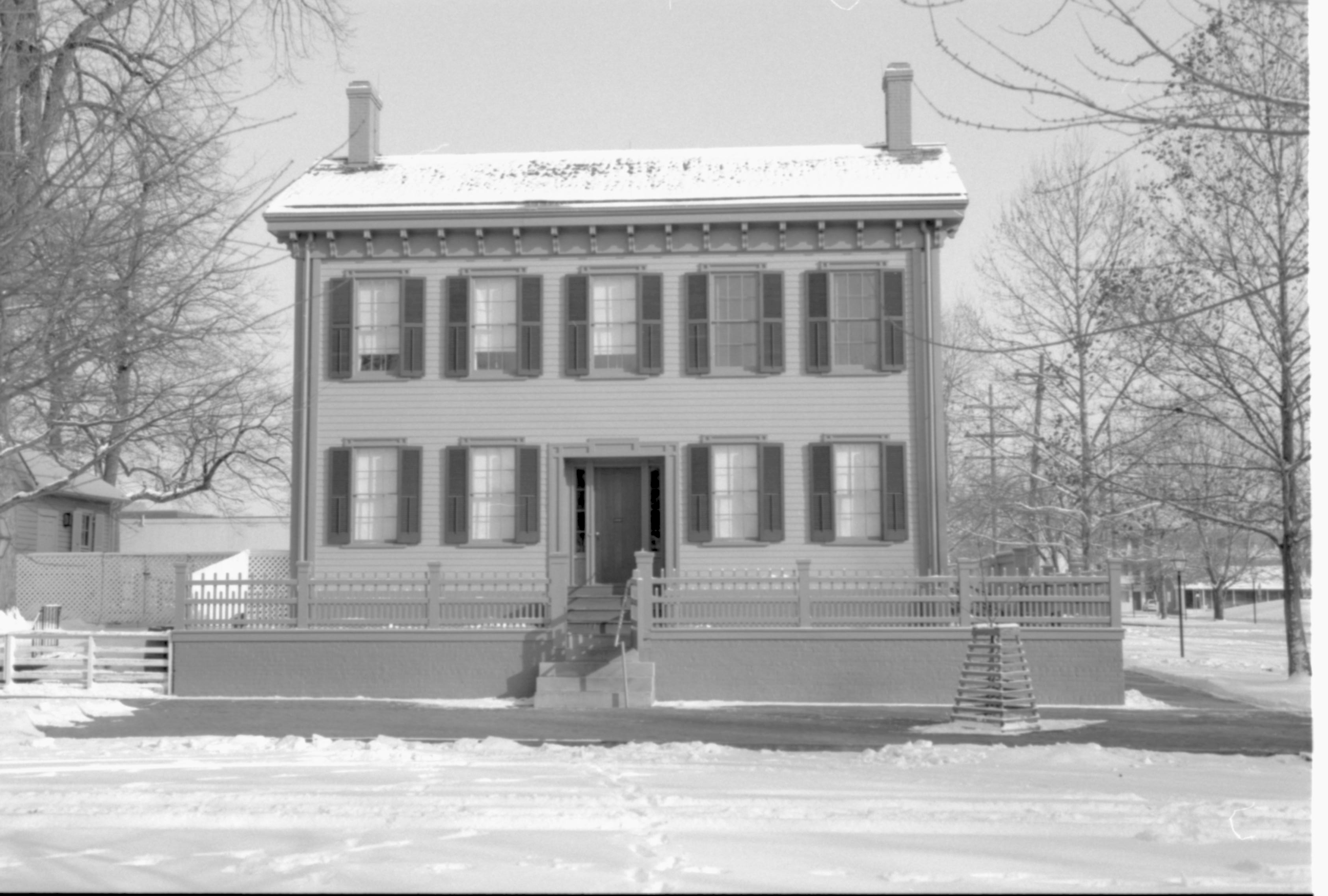 Lincoln Home in snow, cleared brick plaza in front, elm tree in cage on right.  Corneau House on far left, B-2 in background left. Former Travel Lodge Motel in background far right. Maintenance vehicle tracks and footprints in snow in foreground Looking East from boardwalk west of 8th Stret snow, Lincoln Home, Corneau, B-2, Travel Lodge Motel, brick plaza, elm tree, 8th Street