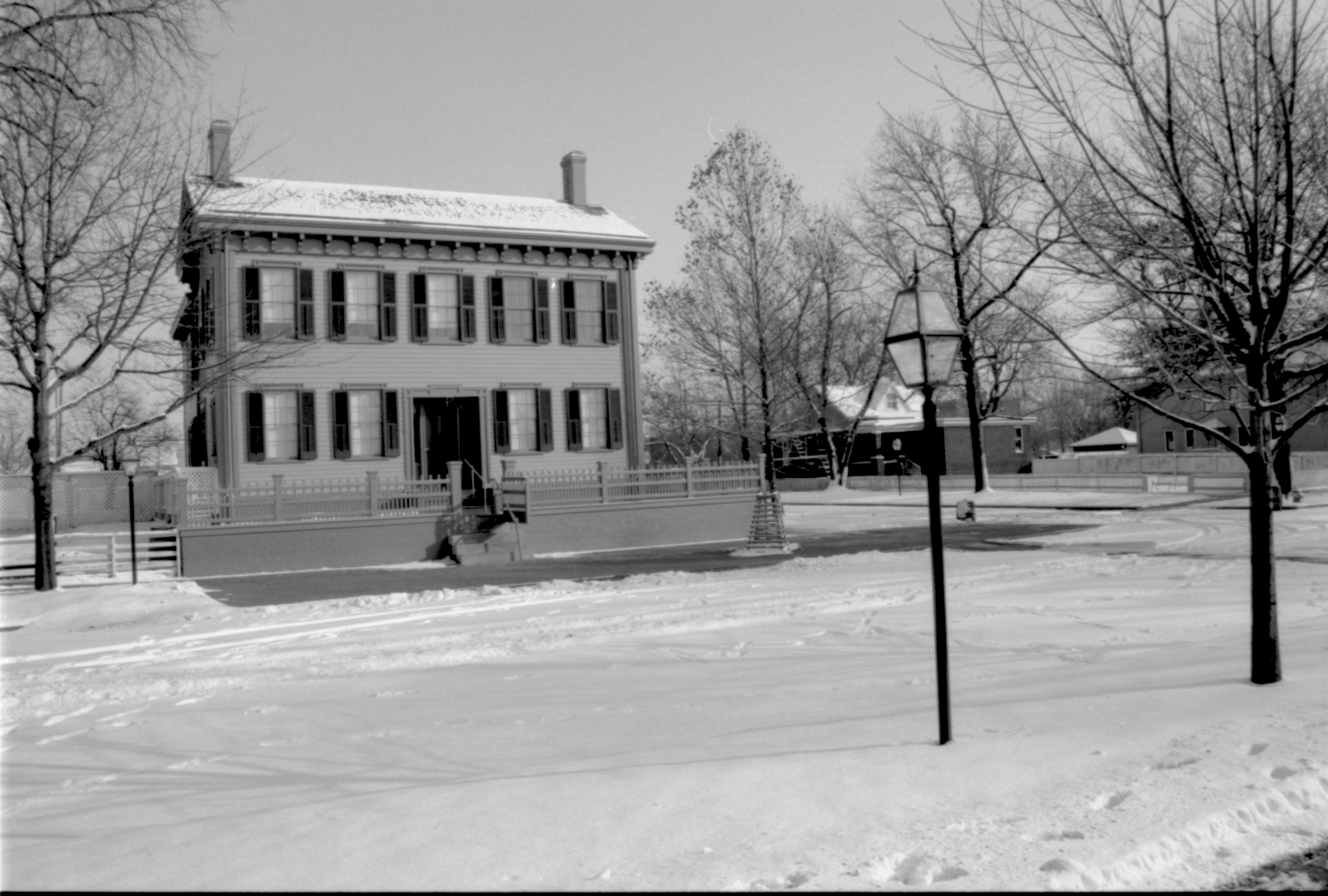 Lincoln Home in snow, cleared brick plaza in front, elm tree in cage on right.  Arnold House in background right, Cook House on far right, Robinson Shed in far background right between Arnold and Cook Houses . Maintenance vehicle tracks and footprints in snow in foreground Looking Southeast from boardwalk west of 8th Street snow, Lincoln Home, Arnold, Cook, Robinson Shed, brick plaza, elm tree, 8th Street