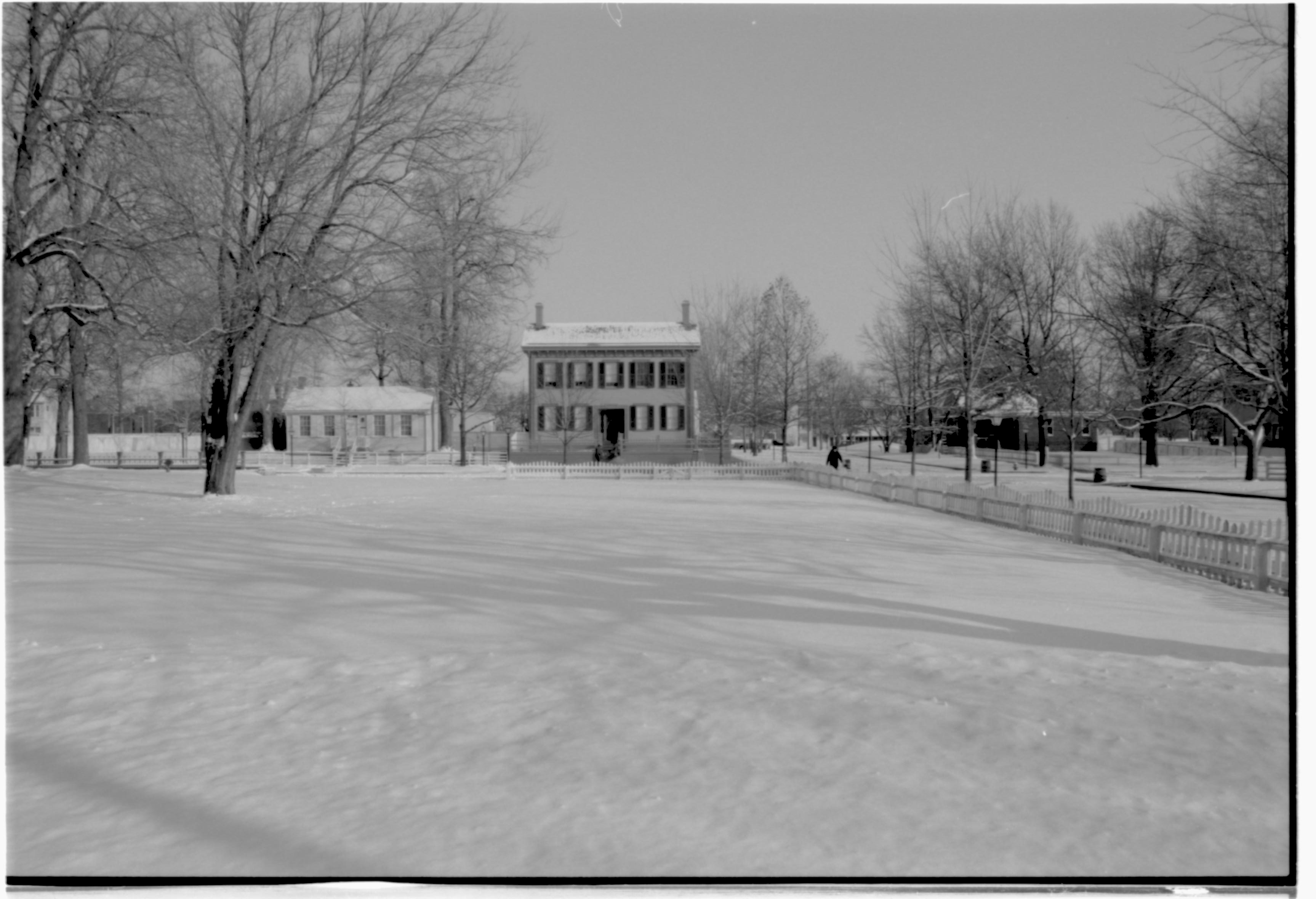 Lincoln neighborhood in the snow.  Lincoln Home in center, Corneau House on left, Morse House in far left background, Arnold House on right. Burch Lot in foreground. Visitor walks on boardwalk along Jackson Street. Looking East from alley behind Visitor Center across Burch Lot, Block 7 Lot 9 snow, Lincoln Home, Morse, Corneau, Arnold, 8th Street, Jackson Street, Burch Lot, visitor