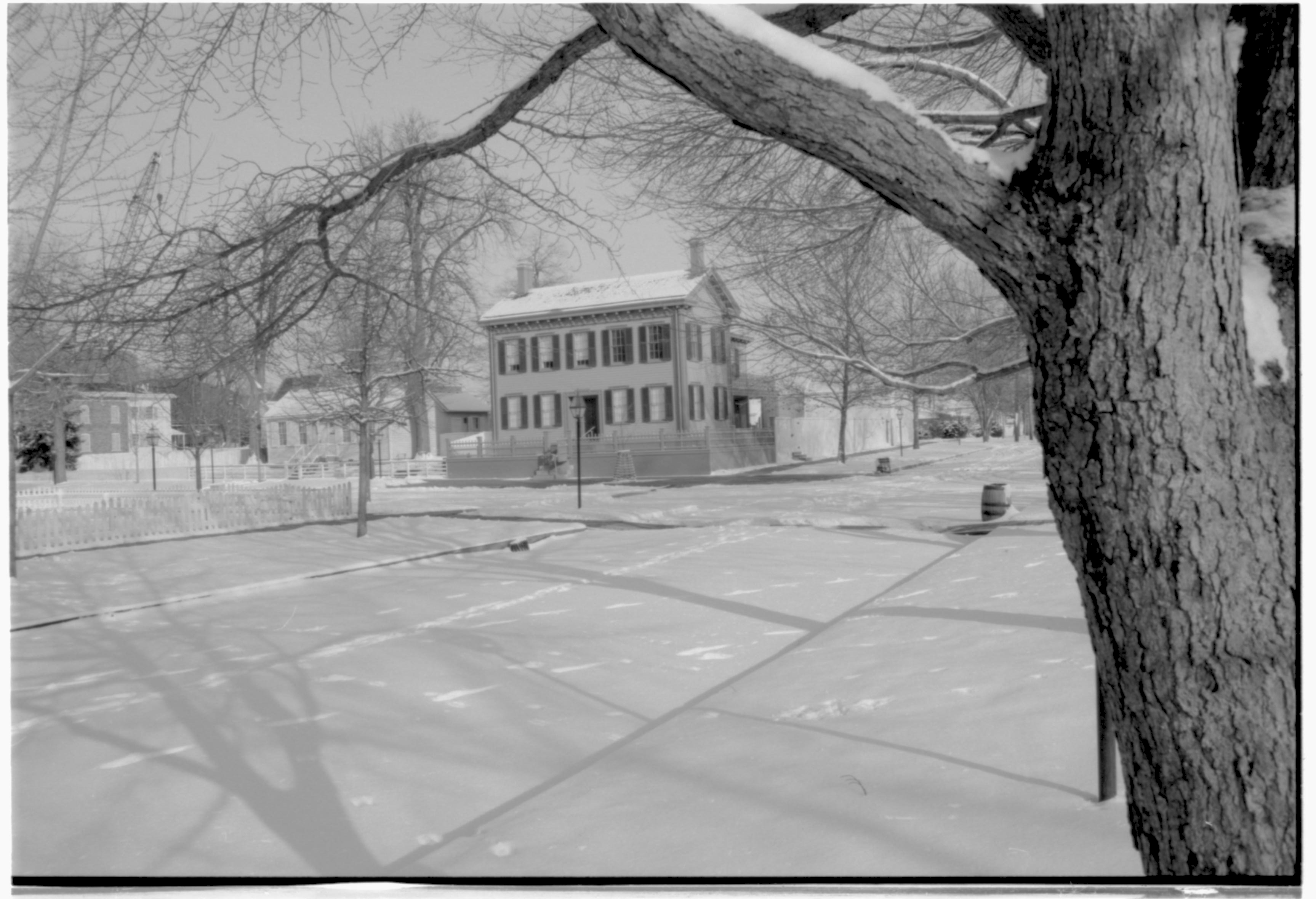 Lincoln neighborhood in snow.  Lincoln Home in center, Corneau House on left, Conference Center on far left, Lincoln Barn visible behind fence along Jackson Street behind Lincoln Home. Red Oak tree in foreground right, footprints in snow throughout. Looking Northeast from alley behind Visitor Center at intersection w/Jackson Street snow, Lincoln Home, Corneau, Lincoln Barn, Conference Center, Jackson Street, Red Oak