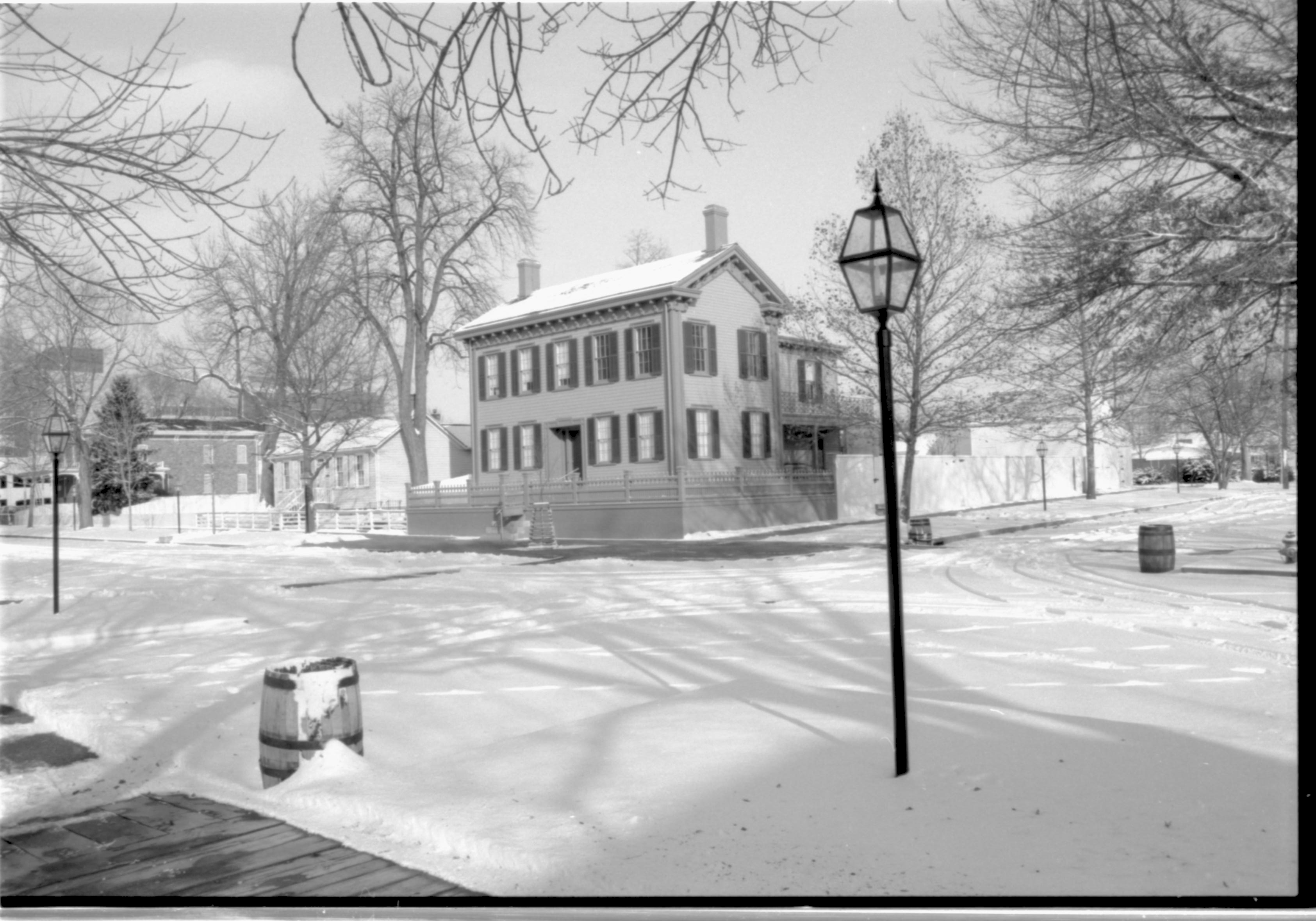 Lincoln Home in the snow, cleared brick plaza in front with elm tree in cage on plaza.  Corneau House on left, Conference Center in brick on far left. Lincoln Barn behind fence along side of Lincoln Home on Jackson Street. Trash barrels visible on corners near street lights and cleared boardwalks. Looking Northeast from just south of 8th and Jackson Street intersection snow, Lincoln Home, Corneau, Conference Center, Lincoln Barn, 8th Street, Jackson Street, boardwalk, fence