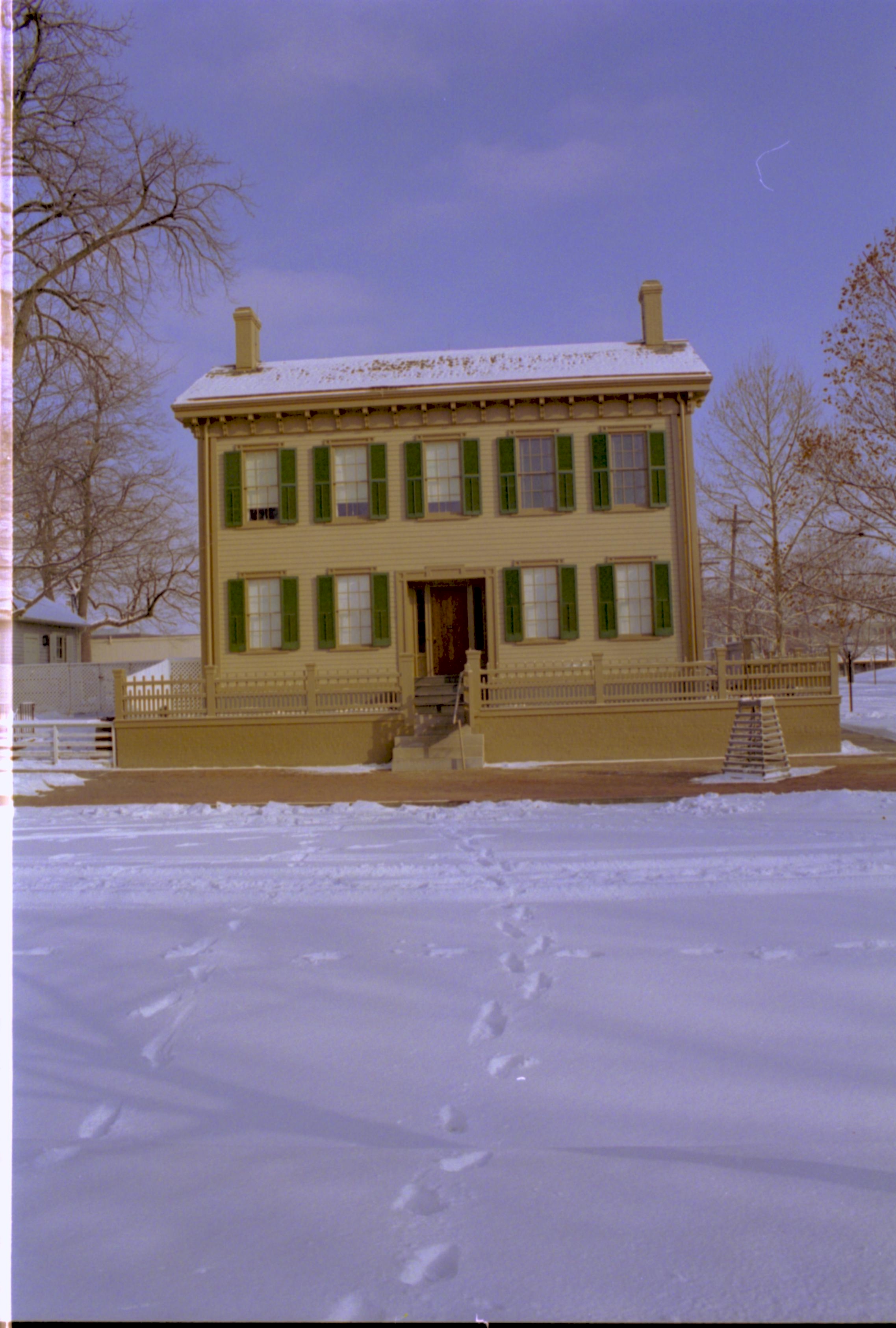 Lincoln Home in snow, cleared brick plaza in front, elm tree in cage on right. Corneau House on left, B-2 in background left. Maintenance vehicle tracks and footprints in snow throughout. Looking East from 8th Street snow, Lincoln Home, Corneau, B-2, 8th Street, brick plaza, elm tree