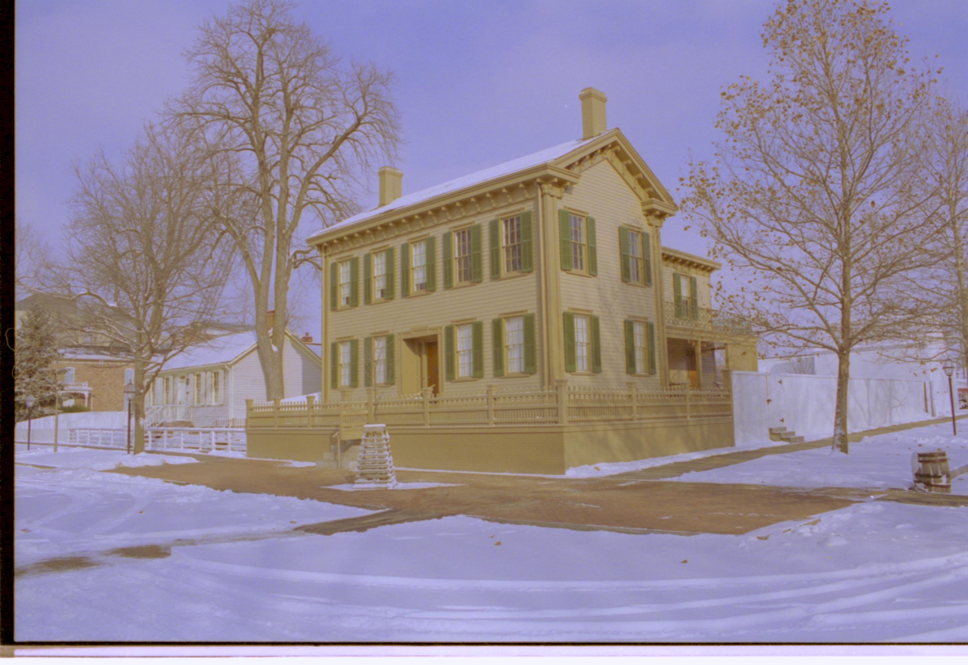 Lincoln Home in snow, cleared brick plaza in front, elm tree in cage on plaza. Corneau House on left, Conference Center in brick on far left. Lincoln Barn and Lincoln Woodshed visible behind fence on far right. Trash barrel in gutter along Jackson Street Looking Northeast from 8th and Jackson Street intersection snow, Lincoln Home, Corneau, Conference Center, Lincoln Barn, Lincoln Woodshed, 8th Street, Jackson Street, brick plaza, elm tree