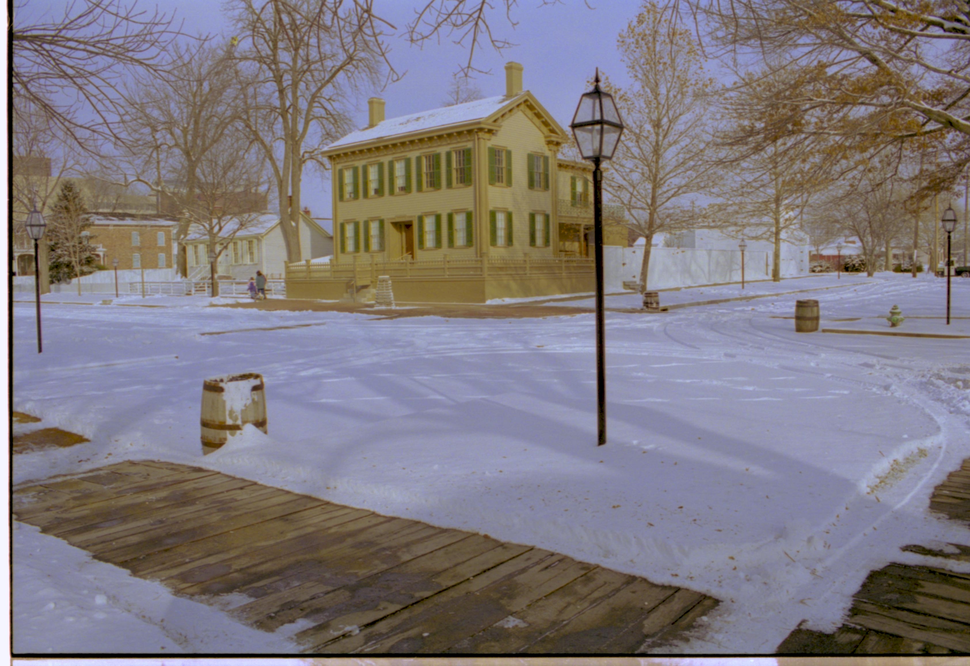 Lincoln neighborhood in snow, Lincoln Home in center, cleared brick plaza in front, elm tree in cage on plaza.  Corneau House on left with visitors walking in front. Conference Center on far left. Lincoln Barn and Lincoln Woodshed behind fence on right. Cleared boardwalks in foreground and along side of Lincoln Home. Trash barrels on three corners. Looking Northeast from just off 8th and Jackson Street intersection snow, Lincoln Home, Corneau, Conference Center, Lincoln Barn, Lincoln Woodshed, 8th Street, Jackson Street