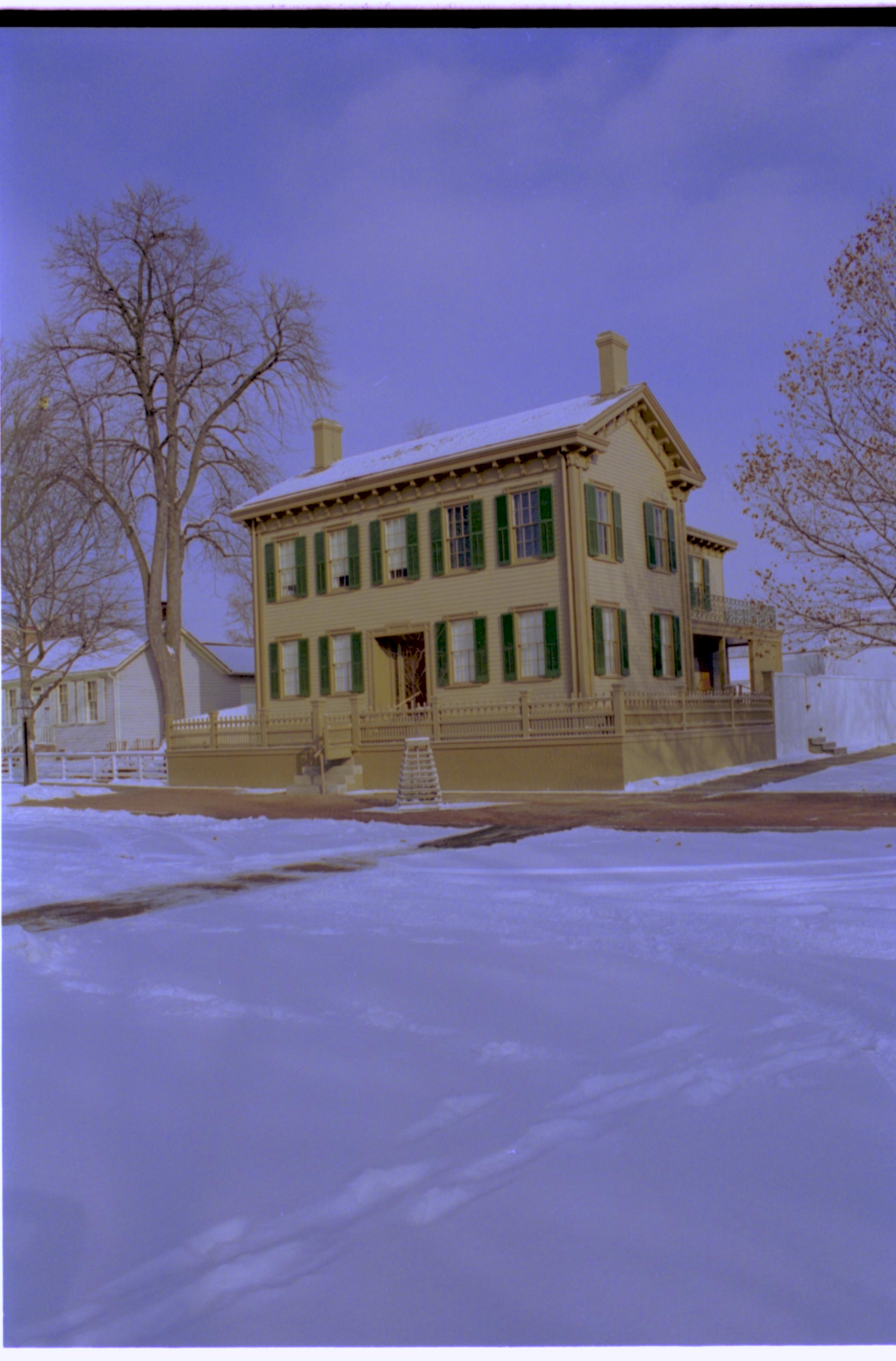 Lincoln Home in snow, cleared brick plaza in front, elm tree in cage on plaza.  Corneau House on left, Lincoln Barn and Lincoln Woodshed behind fence on right. Footprints in snow in foreground. Looking Northeast from 8th and Jackson Street intersection snow, Lincoln Home, Corneau, brick plaza, elm tree, 8th Street, Jackson Street