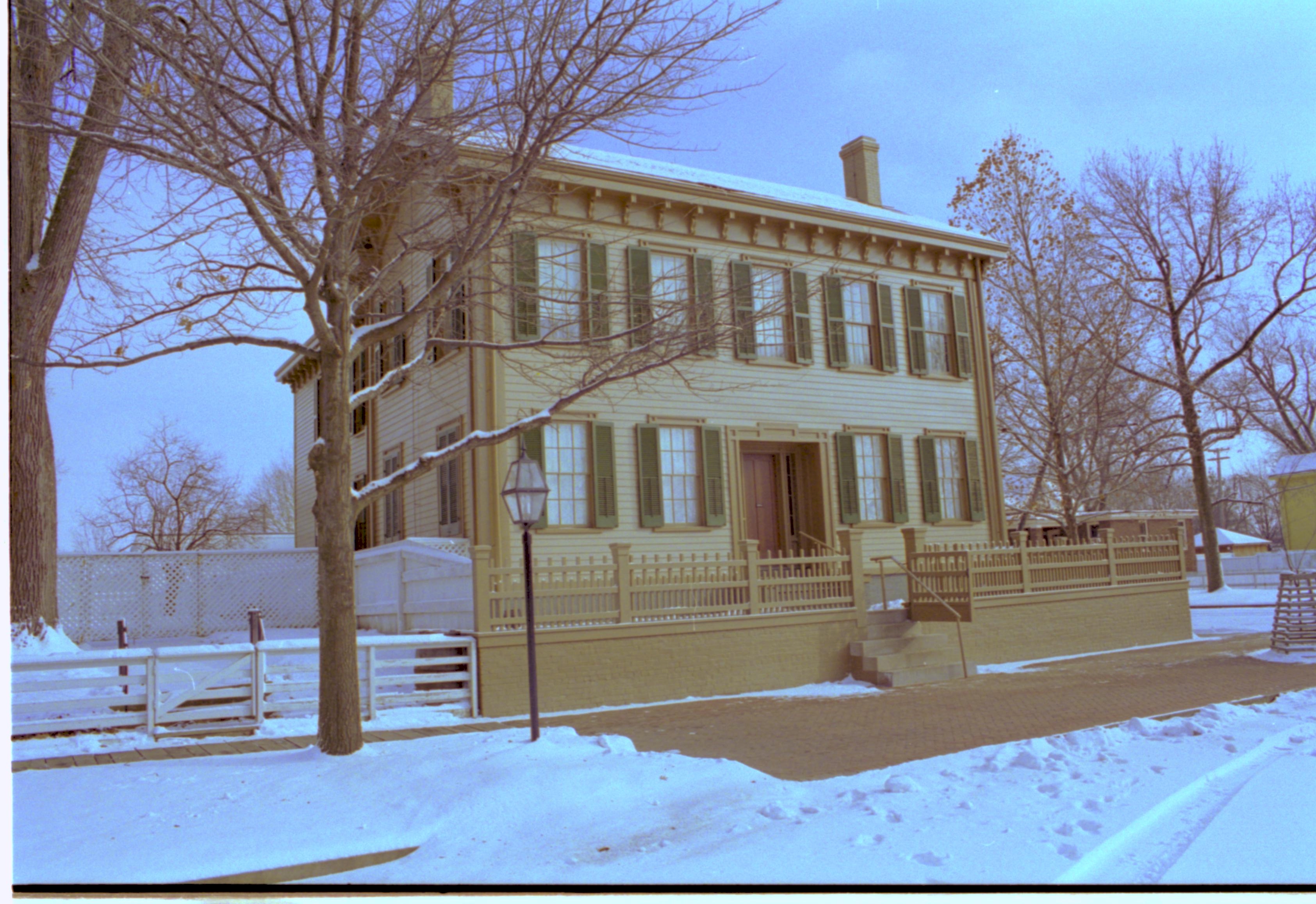 Lincoln Home in snow, cleared brick plaza in front, elm tree in cage on far right. Cook House in yellow on far right background. Maintenance vehicle track and footprints in snow on right. Looking Southeast from 8th Street snow, Lincoln Home, brick plaza, elm tree, Cook, 8th Street