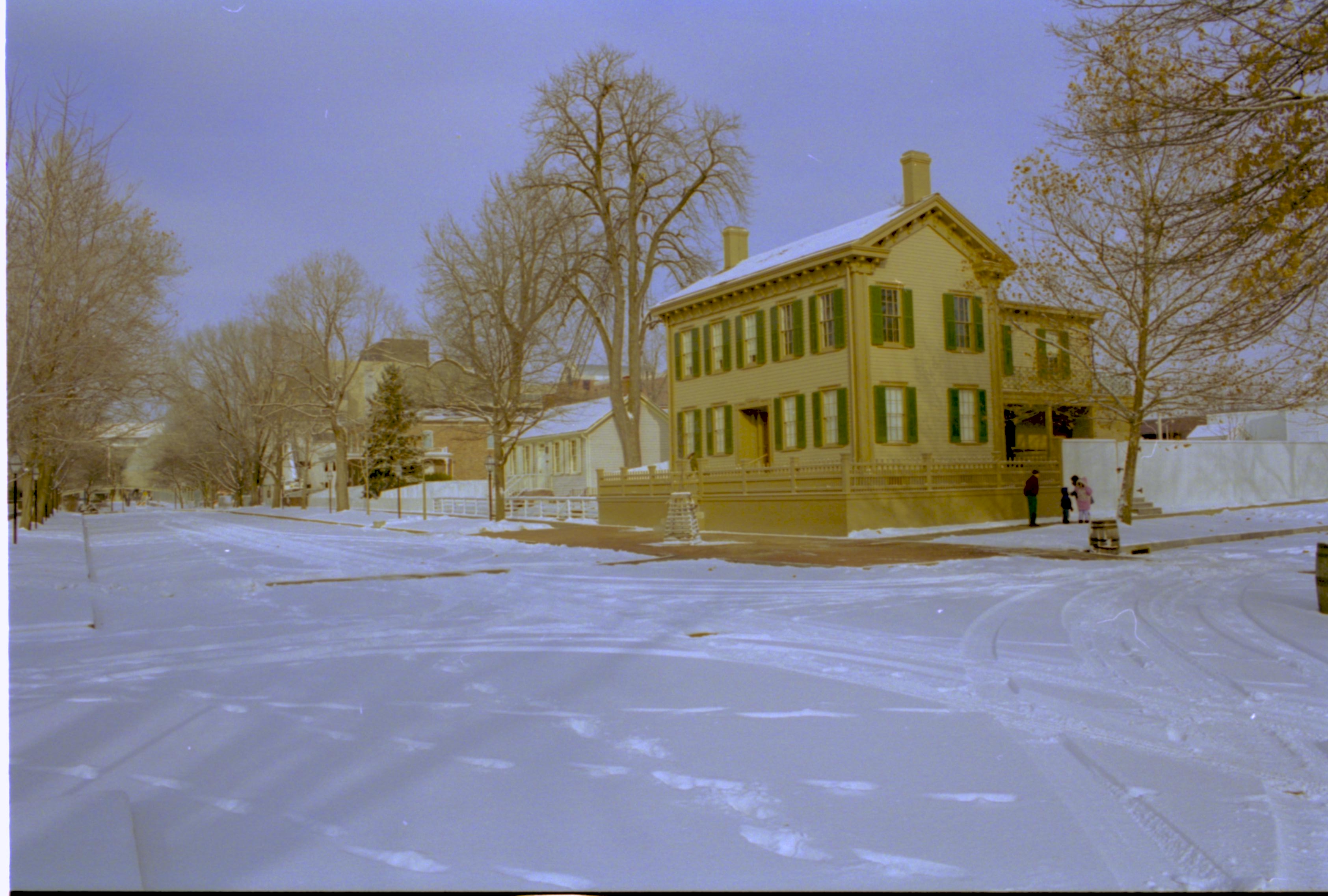 Lincoln neighborhood in snow. Lincoln Home in center with visitors on boardwalk on side of Home.  Corneau House on left, Conference Center in brick in background left. Maintenance vehicle tracks and footprints in snow throughout. Looking North/Northeast from just south of 8th and Jackson Street intersection snow, Lincoln Home, Corneau, Conference Center, 8th Street, Jackson Street, brick plaza, elm tree, visitors