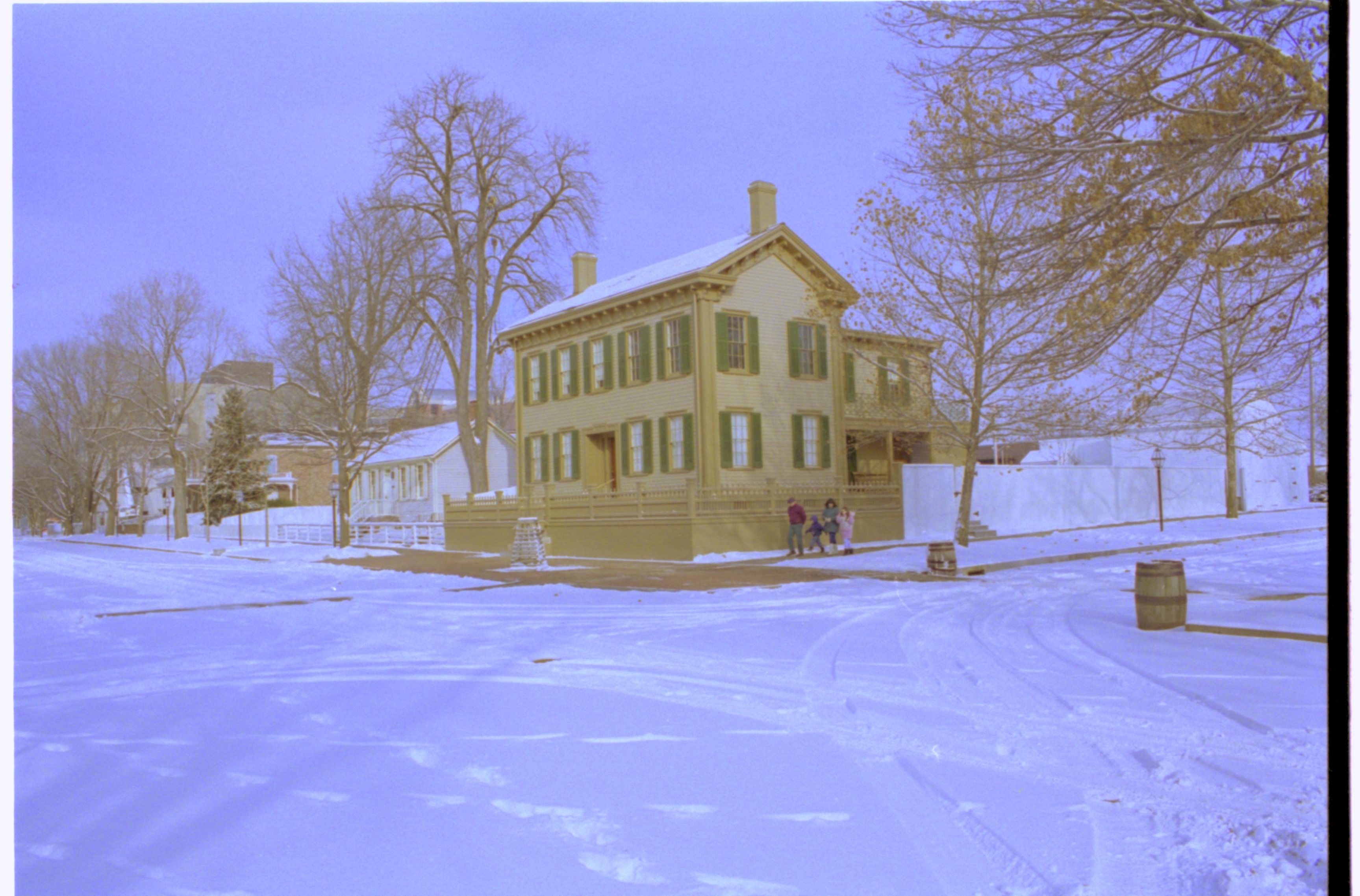 Lincoln neighborhood in snow. Lincoln Home in center with cleared brick plaza in front, elm tree in cage on plaza, visitors on side of house on boardwalk. Corneau House on left, Conference Center in background left. Maintenance vehicle tracks and footprints in snow througout. Looking Northeast from 8th and Jackson Street intersection snow, Lincoln Home, Corneau, Conference Center, brick plaza, elm tree, 8th Street, Jackson Street, visitors