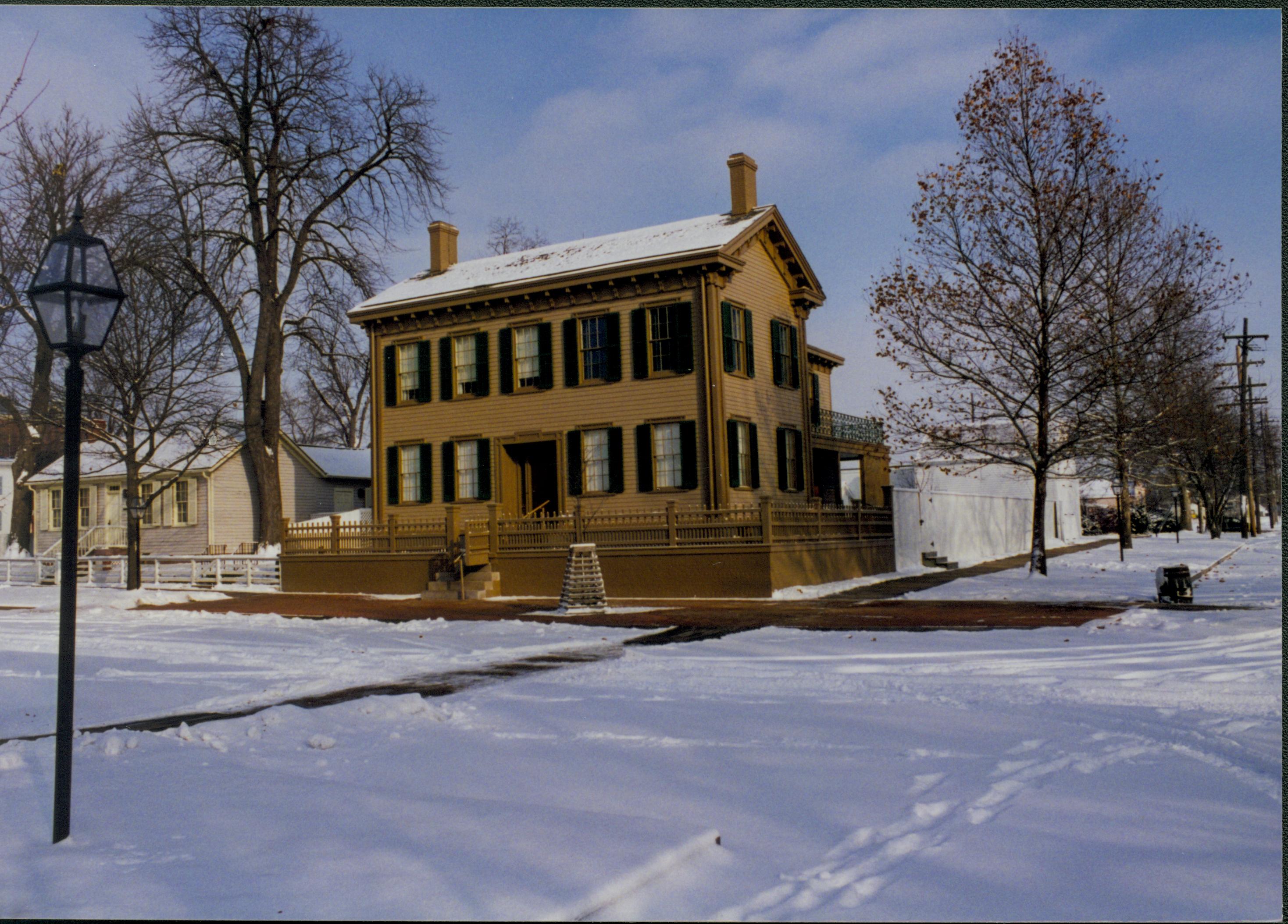 Lincoln neighborhood in the snow from 8th and Jackson Street intersection.  Corneau house on left, brick plaza cleared of snow in front of Lincoln Home, elm tree in cage on plaze.  Trash barrel in gutter along Jackson Street on right.  Footprints and maintenance vehicle tracks visible. Looking Northeast from just west of 8th and Jackson Street intersection snow, Lincoln Home, Corneau, brick plaza, elm tree, 8th Street, Jackson Street