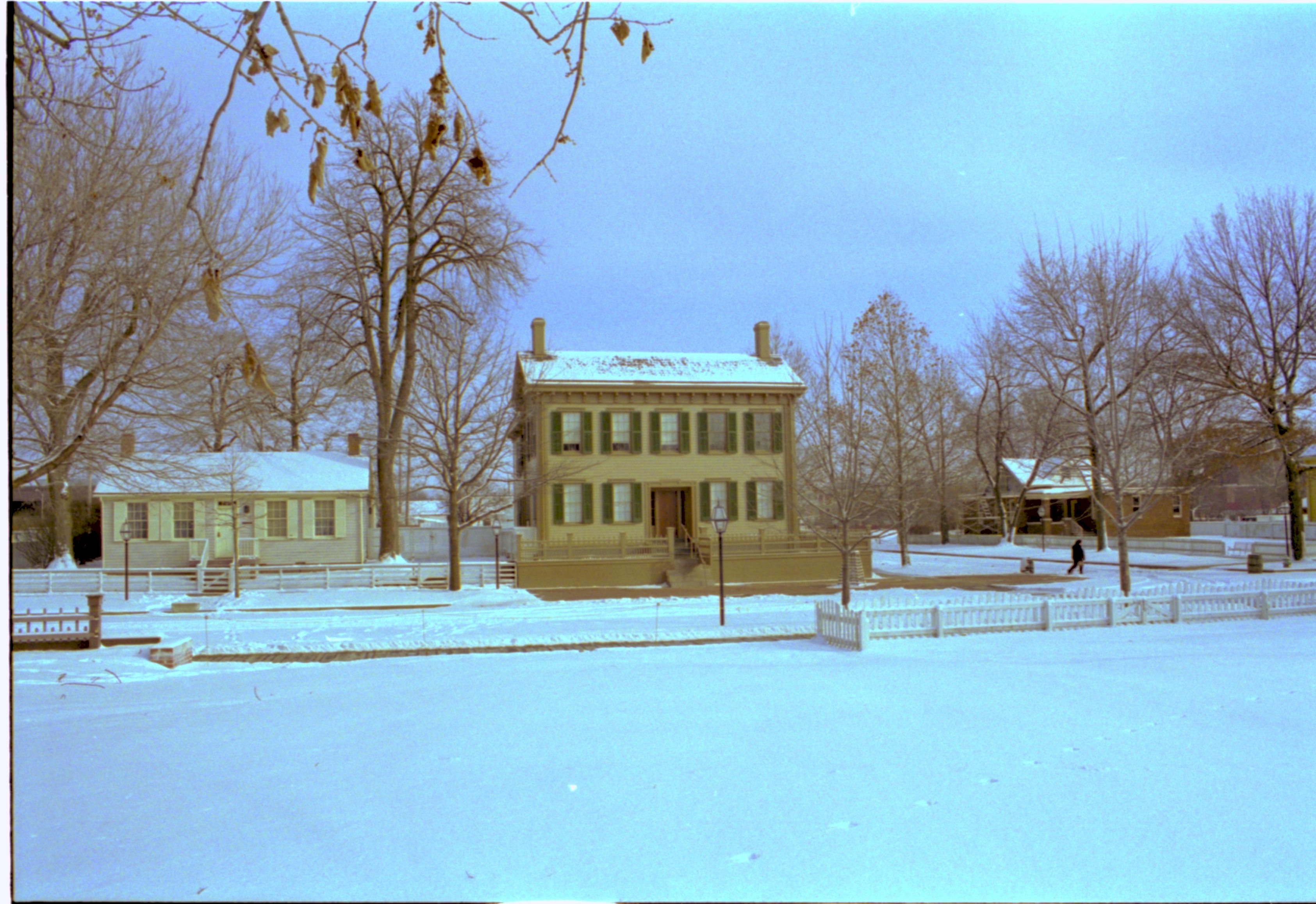 Lincoln neighborhood in snow. Lincoln Home in center with cleared brick plaza in front. Corneau House on left, Arnold House on right. Visitor walking south across Jackson Street. Burch lot (fenced) and Brown lot in foreground Looking East/Southeast from middle of Brown Lot, Block 7, Lot 10 snow, Lincoln Home, Corneau, Arnold House, Brown Lot, Burch Lot, 8th Street