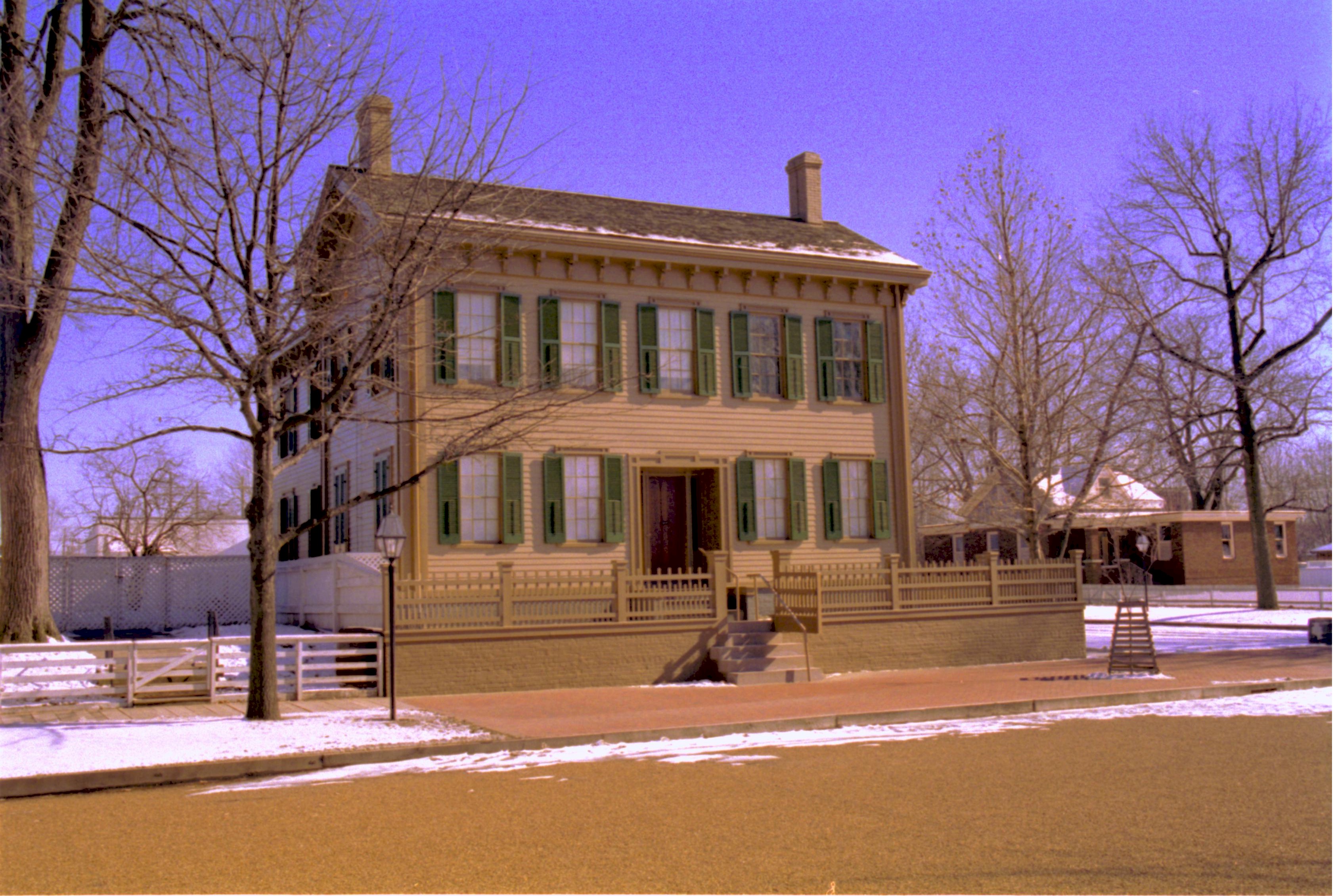 Lincoln Home in winter, scattered snow throughout. Cleared brick plaza in front, elm tree in cage on right. Arnold House in background right behind tree. Lincoln Woodshed in background left behind Home Looking Southeast from 8th Street snow, Lincoln Home, brick plaza, elm tree, Arnold House, 8th Street
