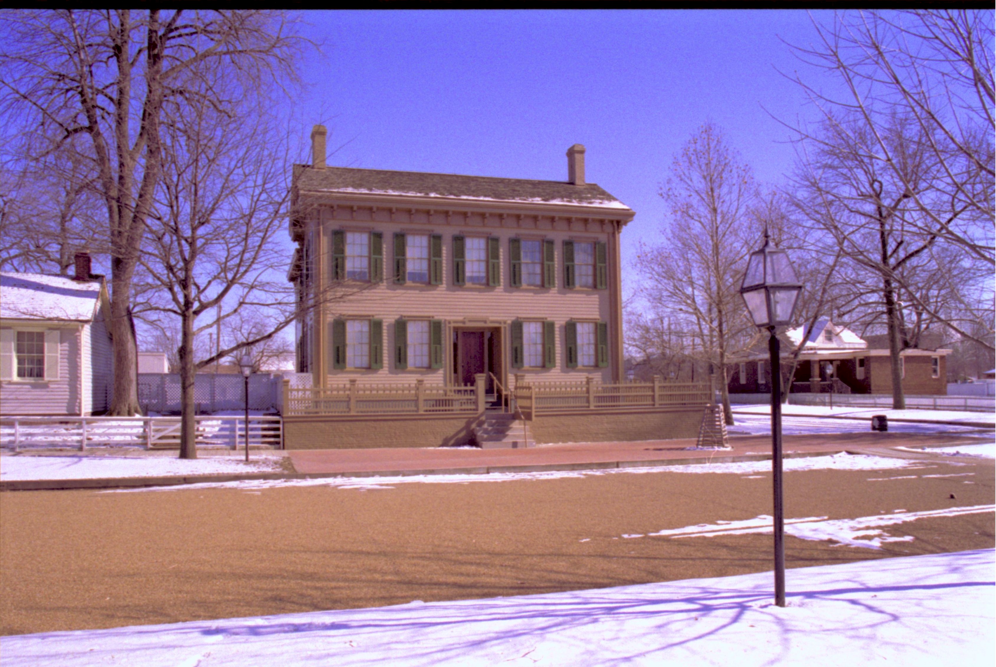 Lincoln Home in winter, snow scattered throughout. Cleared brick plaza in front, elm tree in cage on right. Corneau House on left, Arnold House in backgroun on right Looking East/Southeast from boardwalk west of 8th Street snow, Lincoln Home, Corneau, Arnold House, 8th Street, brick plaza, elm tree