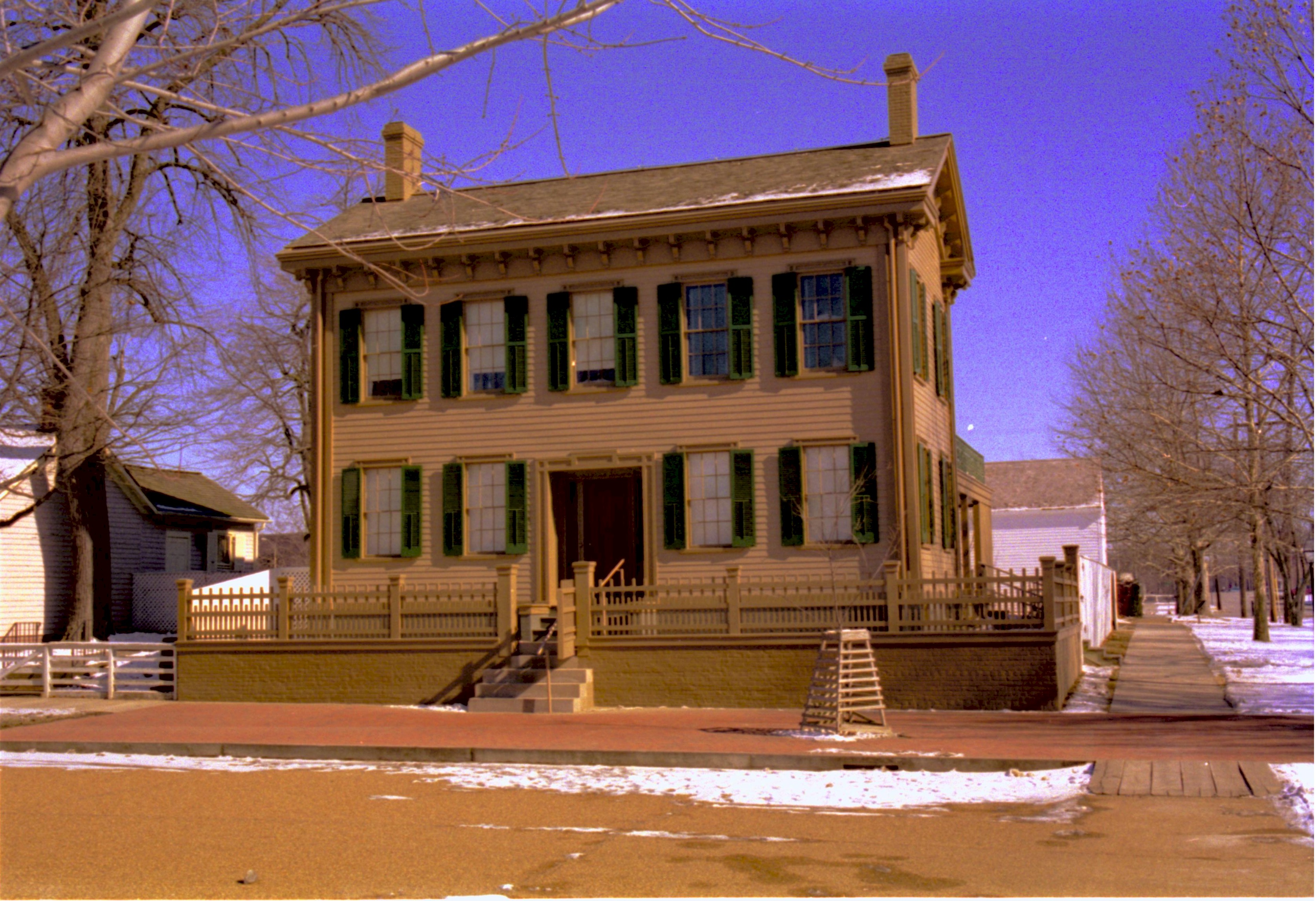 Lincoln Home in winter, snow scattered throughout. Cleared brick plaza in front, elm tree in cage on plaza.  Corneau House on left. Cleared boardwalk along side of hosue. Lincoln Barn in bacground right Looking East from 8th and Jackson Street intersection snow, Lincoln Home, Corneau, Lincoln Barn, 8th Street, Jackson Street, brick plaza, elm tree