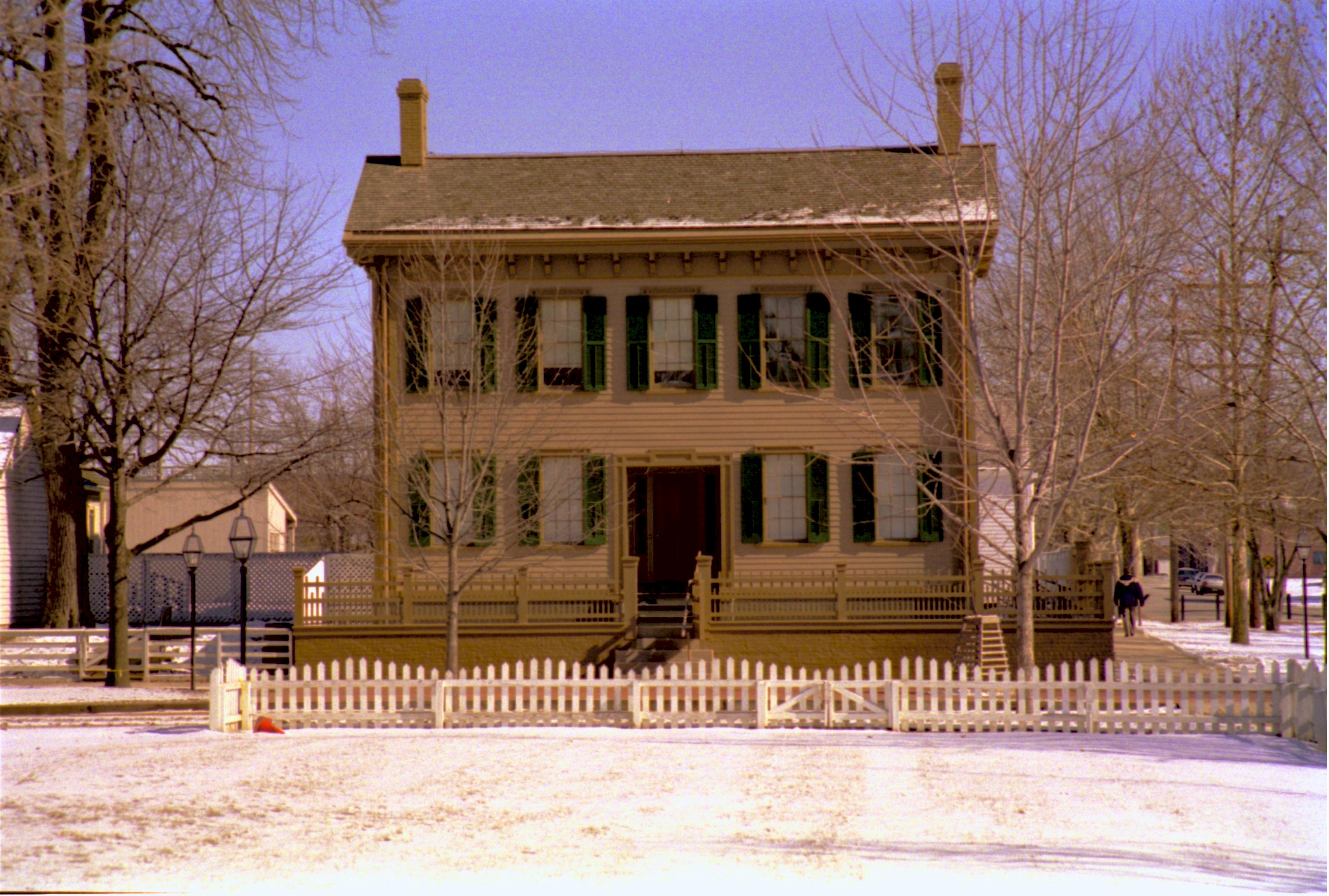 Lincoln Home in winter, snow in foreground on Burch and Brown lots. B-2 in background on left, Corneau House on far left. Visitors walk on boardwalk on side of Lincoln Home on right. Lincoln Barn visible in background behind Home. Looking East from Burch lot, Block 7, Lot 9 snow, Lincoln Home, Corneau, B-2, Lincoln Barn, Burch Lot, Brown Lot, visitors