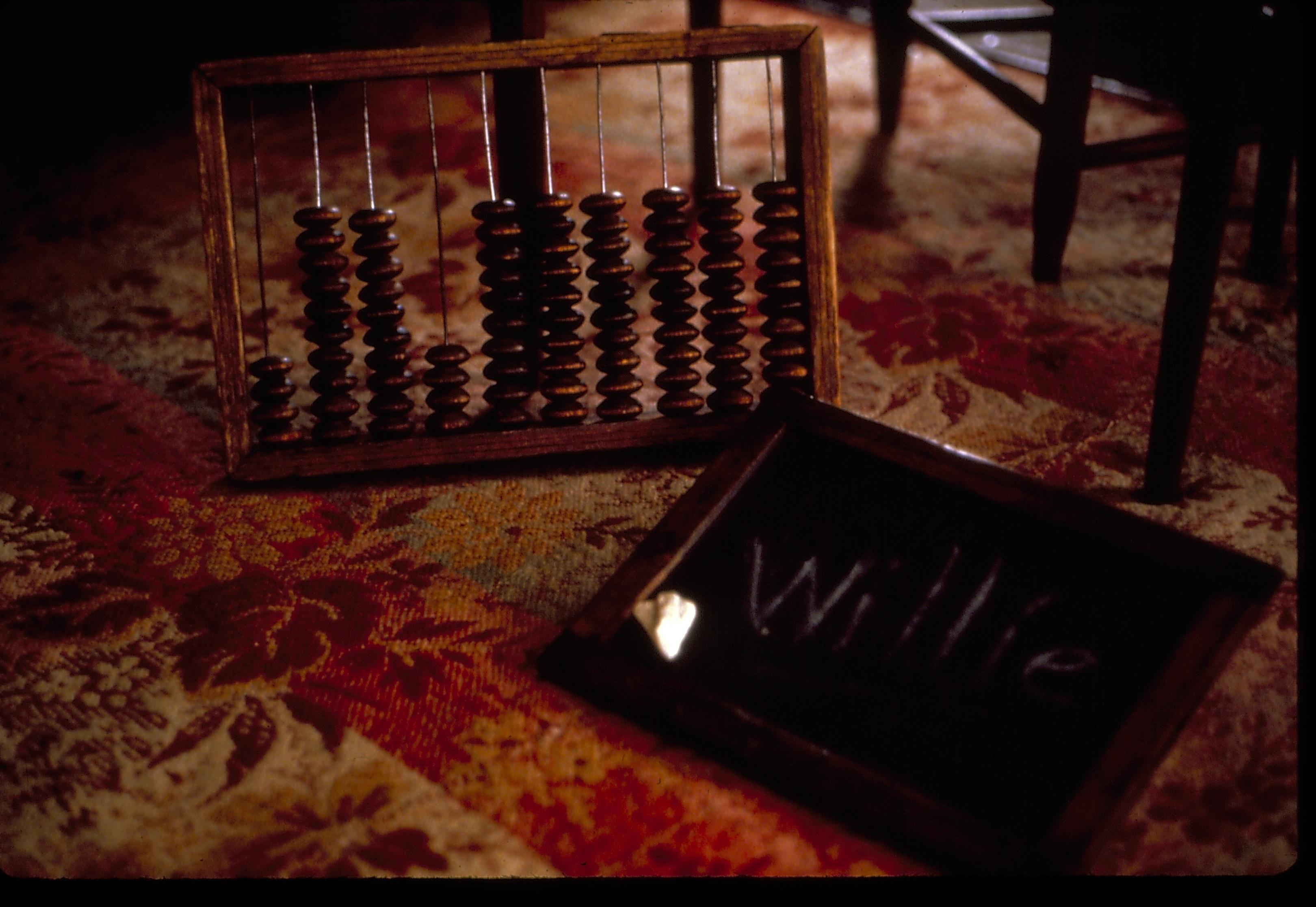 Boys Room (now known as Guest Room) detail of toys on the floor covered in period pattern woven carpet. Features an abacus and chalkboard with "Willie" written on it, chairs in background. Carpet is red and white striped with dark brown floral motif over it.