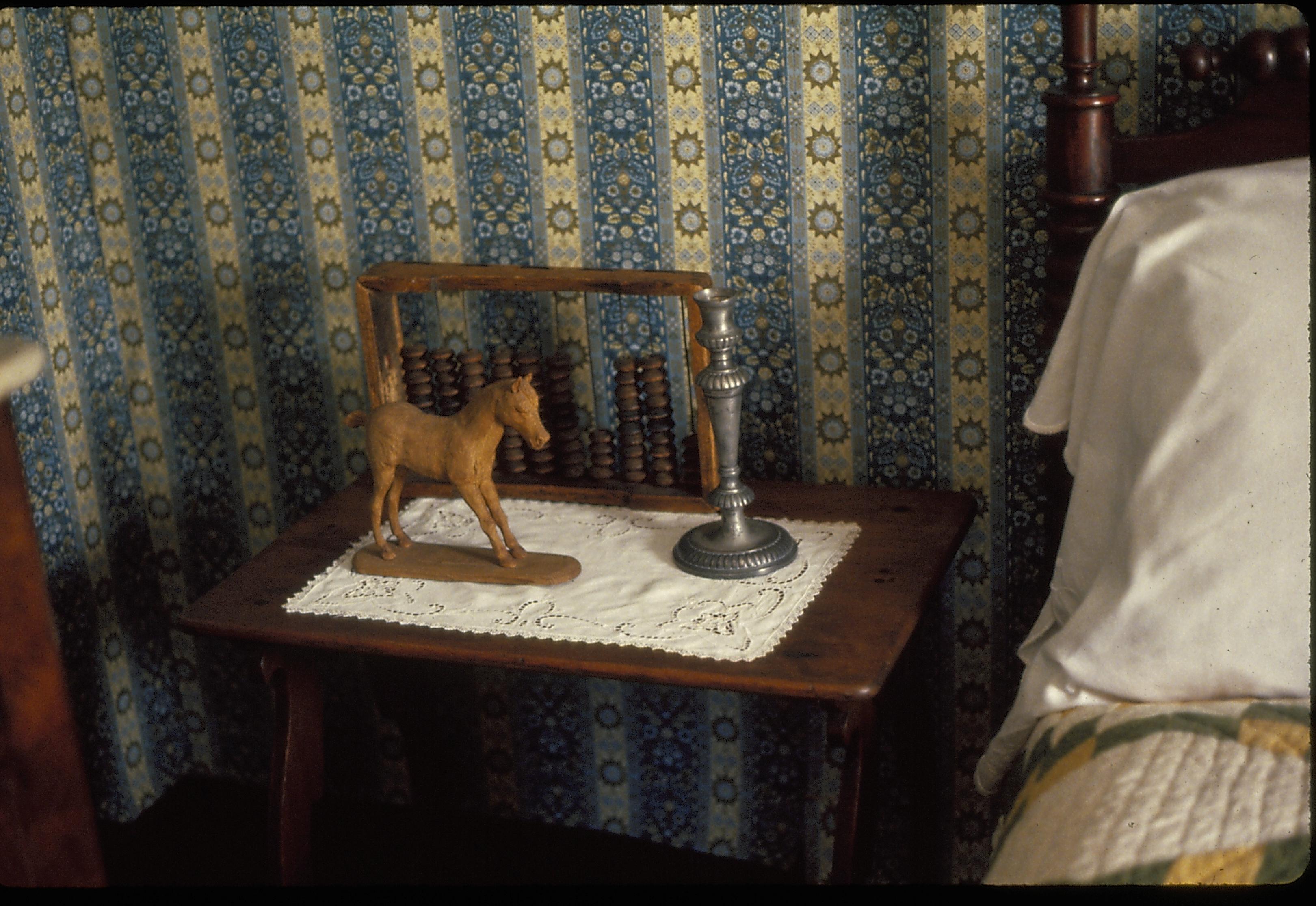 Boys Room, showing bedside table with a wooden horse toy, and abacus, and a pewter candlestick on a doily next to a bed with a white pillow and a green and yellow quilt. Heavily figured blue, yellow and white wallpaper on the walls