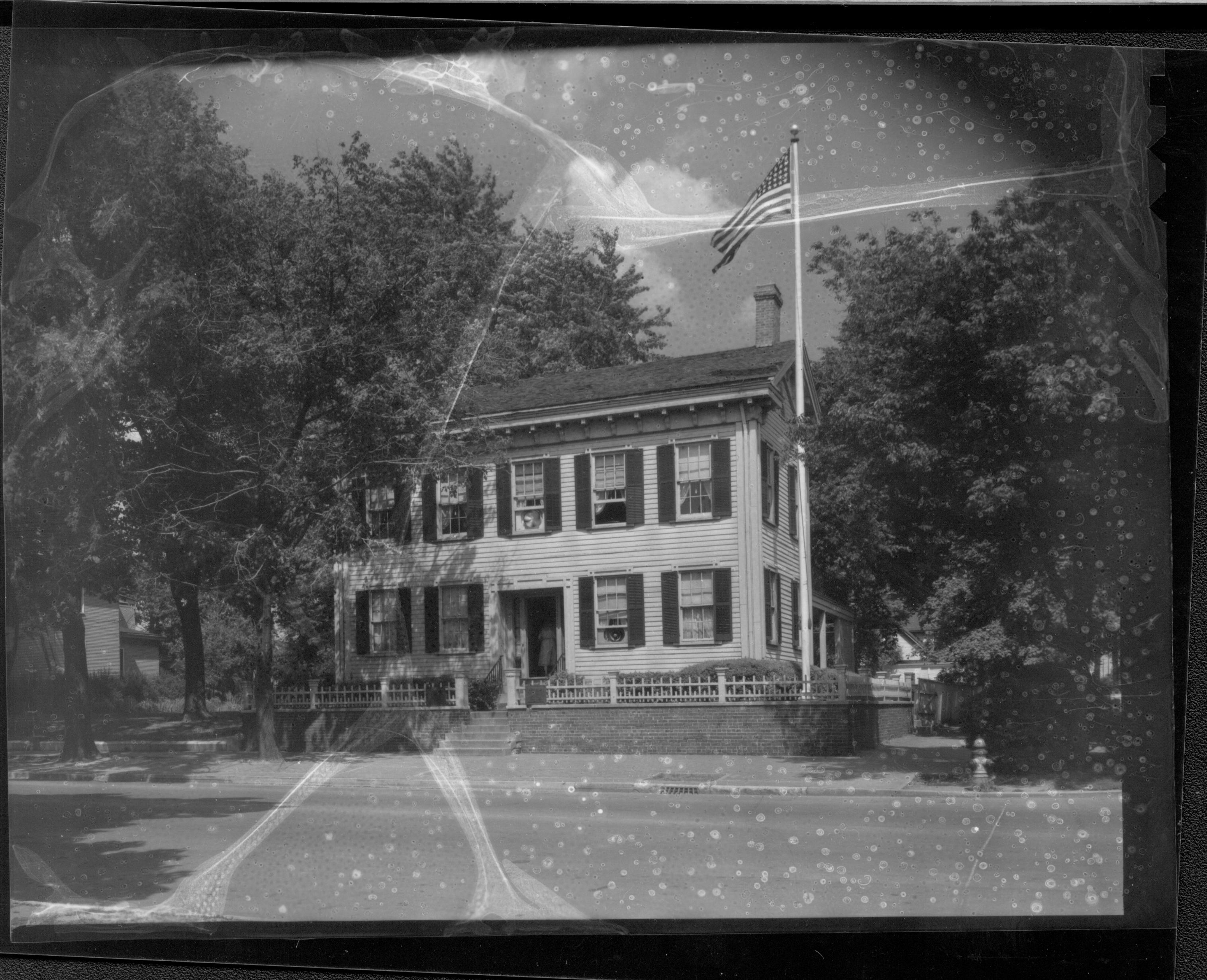 Lincoln Home west (front) elevation in summer surrounded by trees. Flag pole behind picket fence in corner, fire hydrant near street on corner.  Two story house on Bugg lot (Block 10, lot 5) on far left. House visible in far background behind Lincoln Home on Kercheval lot (Block 10, lot 9). Paved street. House appears to be painted white. Looking Northeast from 8th and Jackson Street intersection. Negative deteriorating. Lincoln Home, flagpole, fire hydrant, paved street, painted white, 8th Street, Bugg, Kercheval