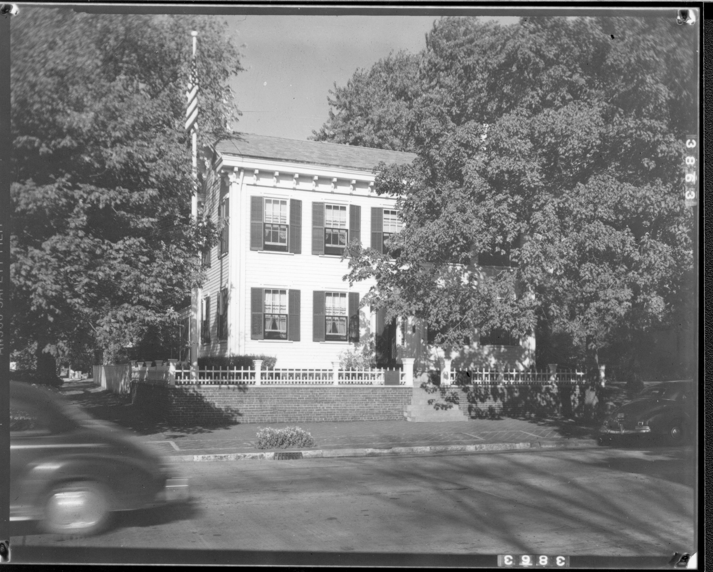 Lincoln Home (reverse image) west (front) elevation in summer surrounded by trees as car drives past on 8th Street. and another with visitor is parked in front on right.  Windows are open with black screens in front. Paved street. House appears to be painted white. Looking East from 8th and Jackson Street intersection Lincoln Home, 8th Street, painted white, cars, visitors, summer
