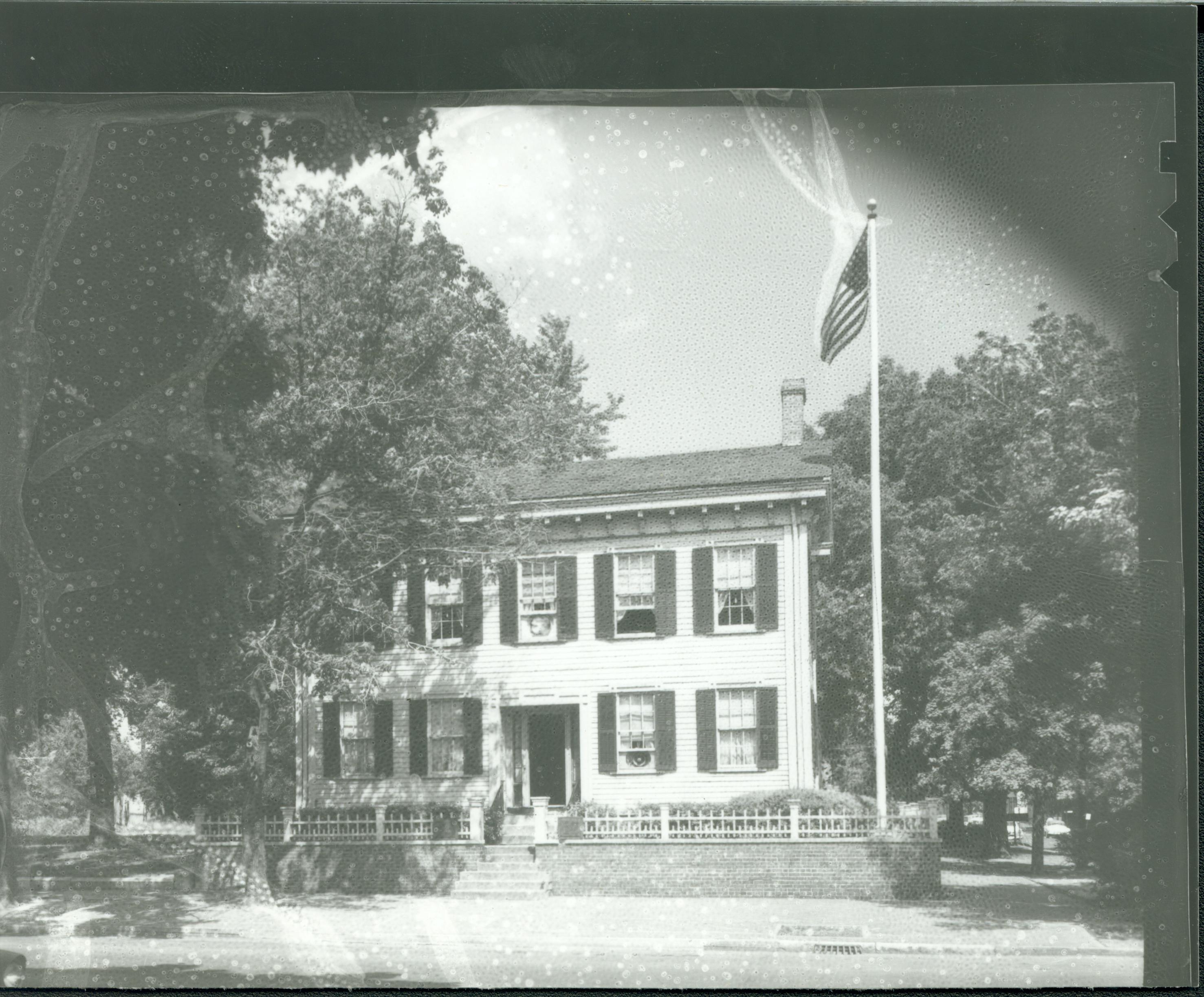 Lincoln Home west (front) elevation in summer surrounded by trees, flagpole in corner behind picket fence. Windows are open without screens, a box fan is visible in upper center window and lower right window. Looking East from 8th Street. Negative deteriorating Lincoln Home, summer, flagpole, 8th Street