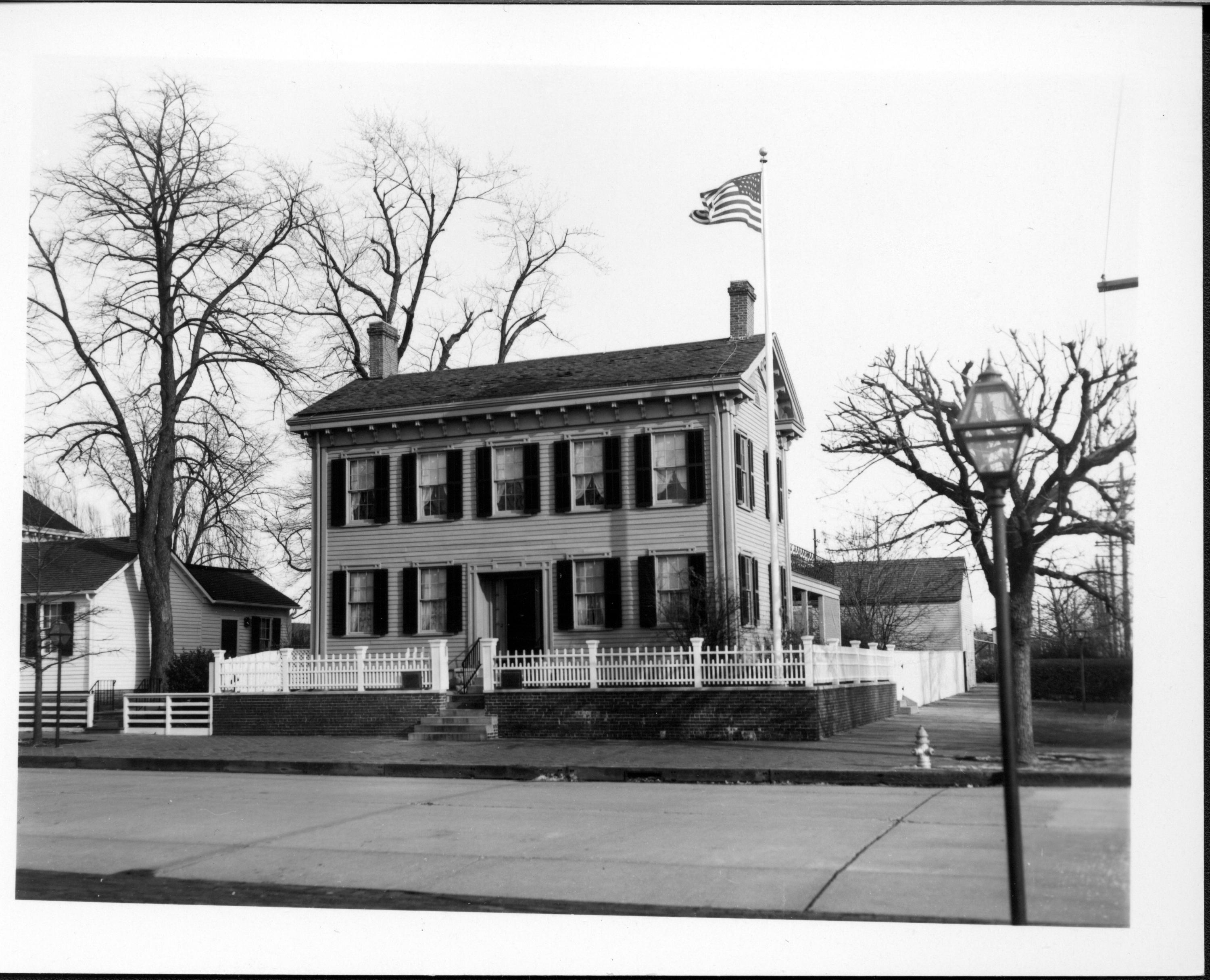 Lincoln Home west (front) elevation with flag pole on corner behind white picket fence. Lincoln Barn visible behind on right. Corneau House visible on left with two-story home on Bugg Lot (Block 10, Lot 5) on far left. Paved street, fire hydrant on corner near street. Square commemorative plaques visible on either side of gate Looking Northeasat from west of 8th Street. Lincoln Home, Corneau, Bugg, Lincoln Barn, flagpole, fire hydrant, 8th Street