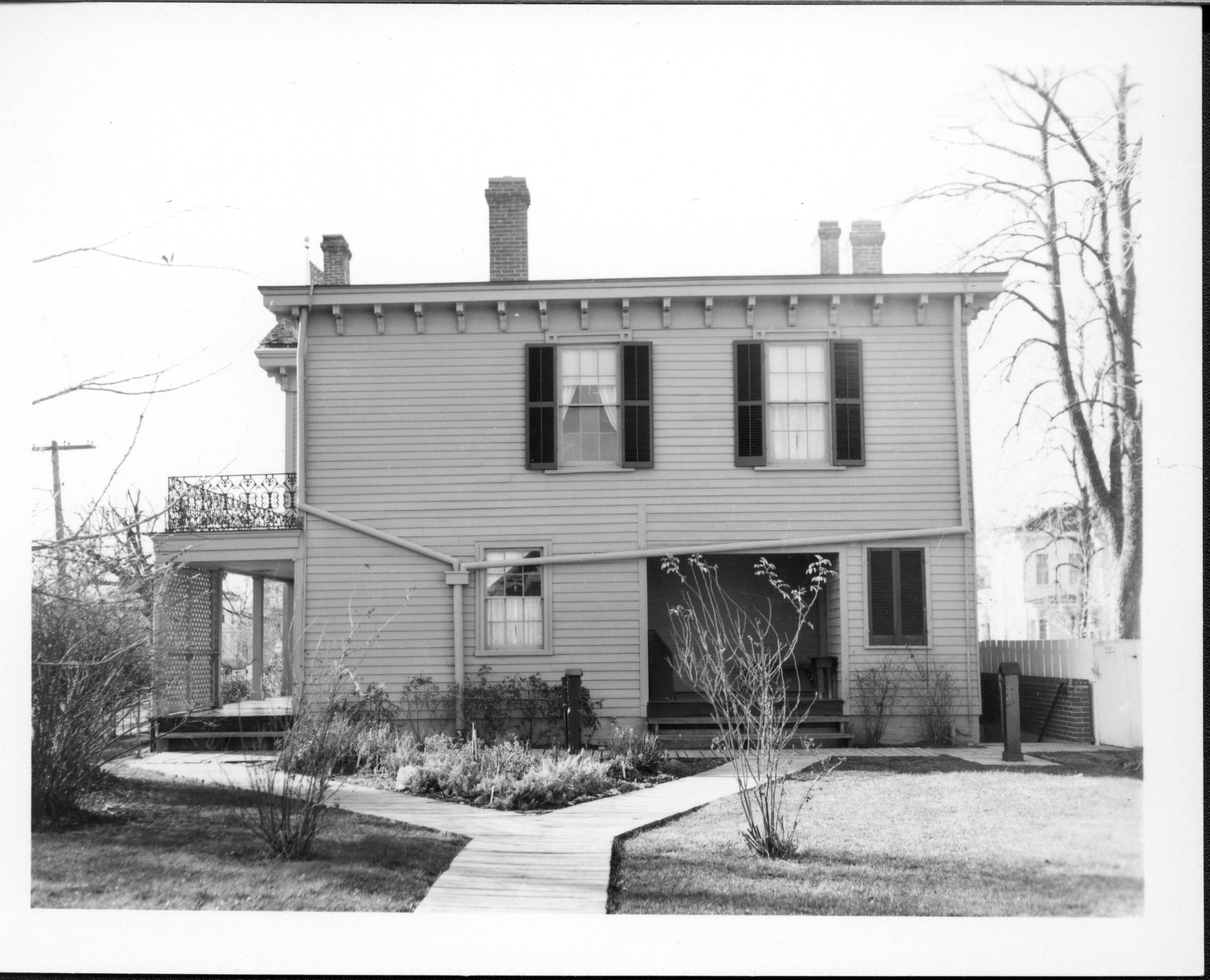 Lincoln Home east (rear) elevation and backyard.  Historically accurate guttering visible across siding and window. Well and cistern pumps seen near sidewalks.  Herb garden is located in triangle near Home, with other bushes scattered around yard.  A stairwell heading to basement seen a right.  Dean House visible in background right. Utility pole seen at left. Looking west from near Lincoln Barn Lincoln Home, backyard, pumps, herb garden, gutters, Dean, utility pole