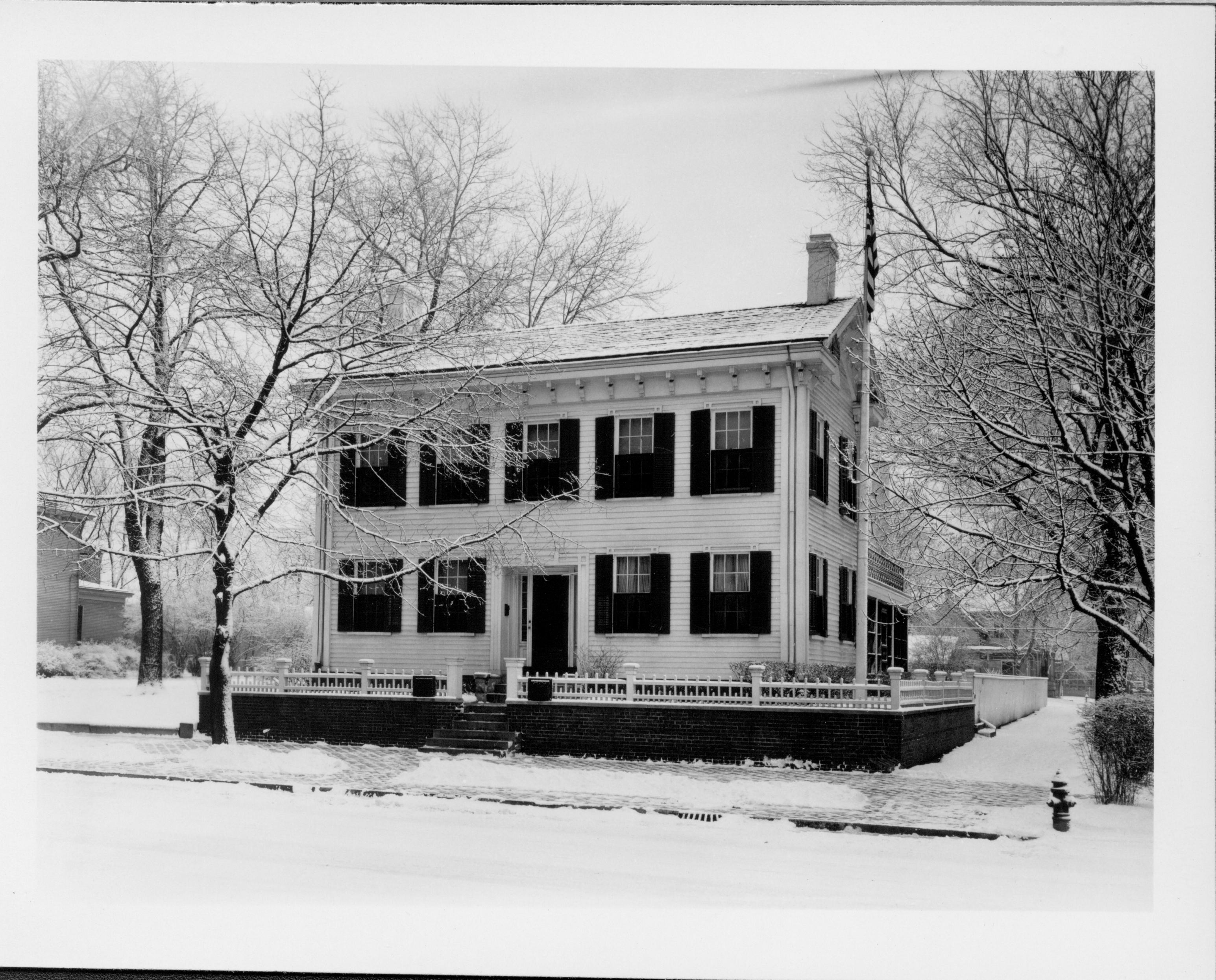 Lincoln Home west (front) elevation in winter, with flagpole on corner behind picket fence.  Fire hydrant visible on corner of brick plaza.  Two story houses visible on Bugg lot (Block 10, Lot 5) on left, and behind Home on Kercheval lot (Block 10, lot 9) in background right.  Back porch is screened in.  Looking Northeast from 8th Street Lincoln Home, 8th Street, snow, Bugg, Kercheval, flagpole, screened back porch