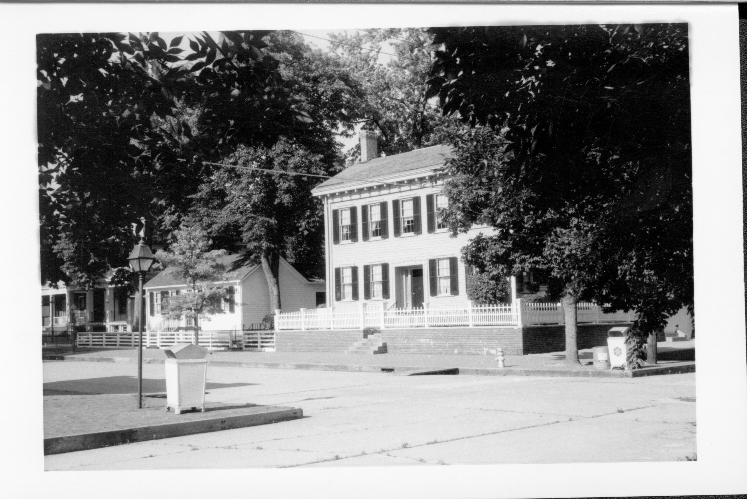Lincoln Home and neighborhood in summer. Lincoln Home west (front) elevation behind closed gate.  Corneau House on left with other houses visible on Bugg (Block 10, Lot 5) and Niles (Block 10, Lot 4) lots.  Trash cans and street light on corners, fire hydrant near street by Lincoln Home.  Paved 8th and Jackson Streets. Looking Northeast from just off 8th and Jackson Street intersections. Lincoln Home, Corneau, Bugg, Niles, 8th Street, Jackson Street, fire hydrant, trash cans, painted white