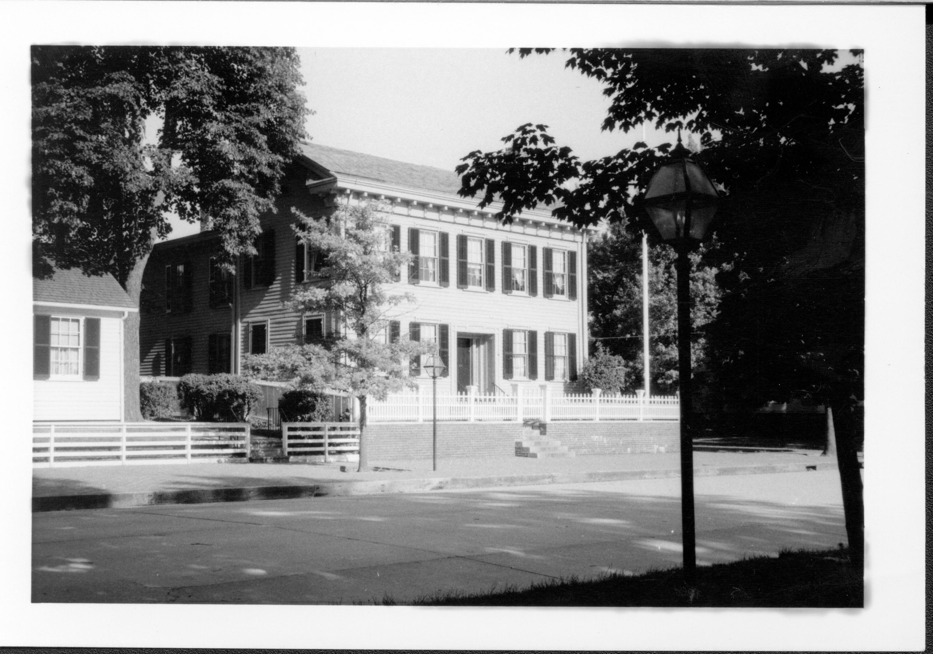 Lincoln Home west (front) elevation behind closed gate. Flagpole on corner behind picket fence on right. Corneau House on left. Street light in foreground. Paved 8th Street. House appears to be painted white. Looking Southeast from west of 8th Street Lincoln Home, Corneau, 8th Street, flagpole, summer, painted white