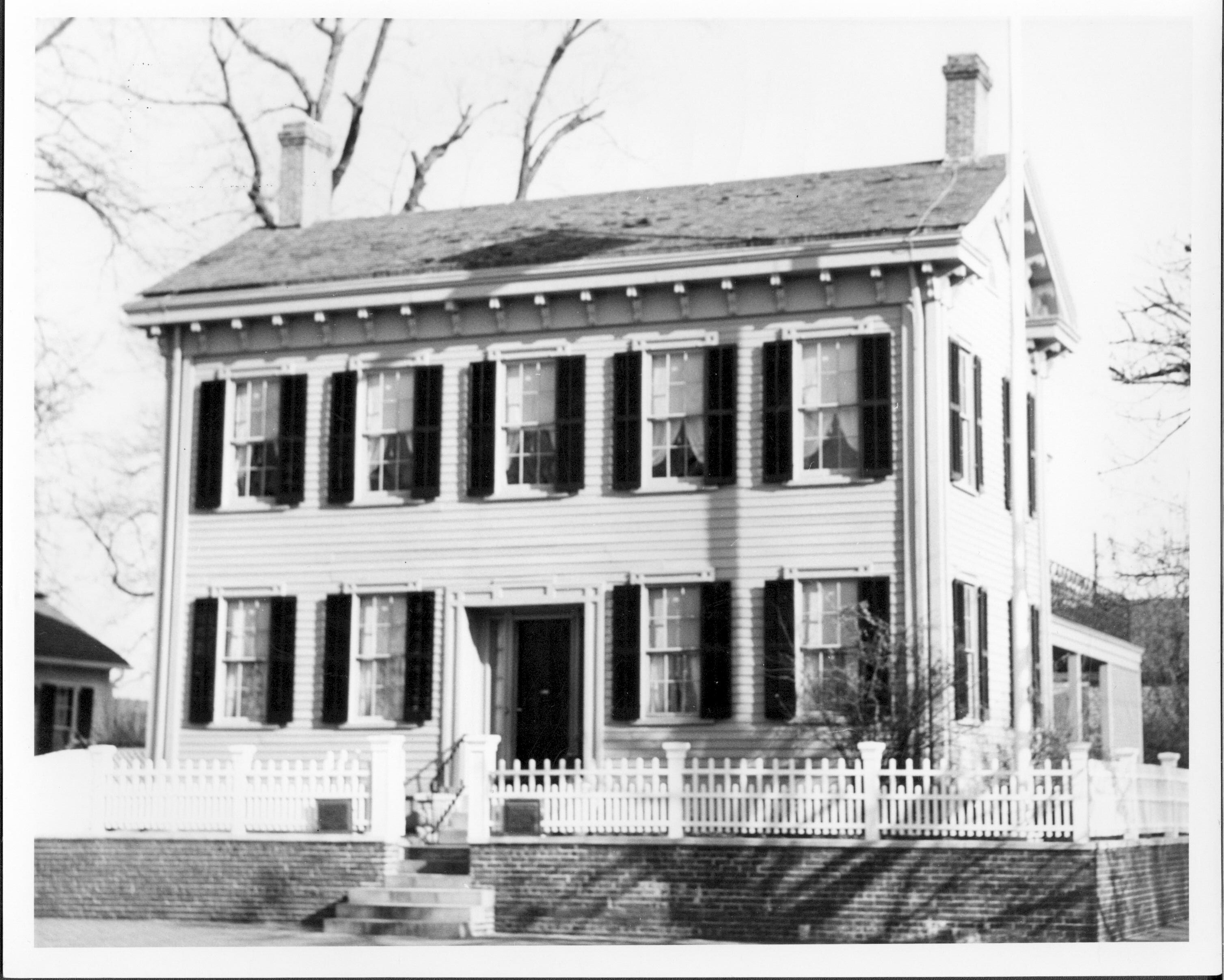 Lincoln Home west (front) elevation. Corneau House on far left. Lincoln Barn just visible behind Lincoln Home on far right.  Square commemorative plaques on either side of open gate. House appears to be painted white. Looking East/Northeast from 8th Street Lincoln Home, Corneau, Lincoln Barn, painted white