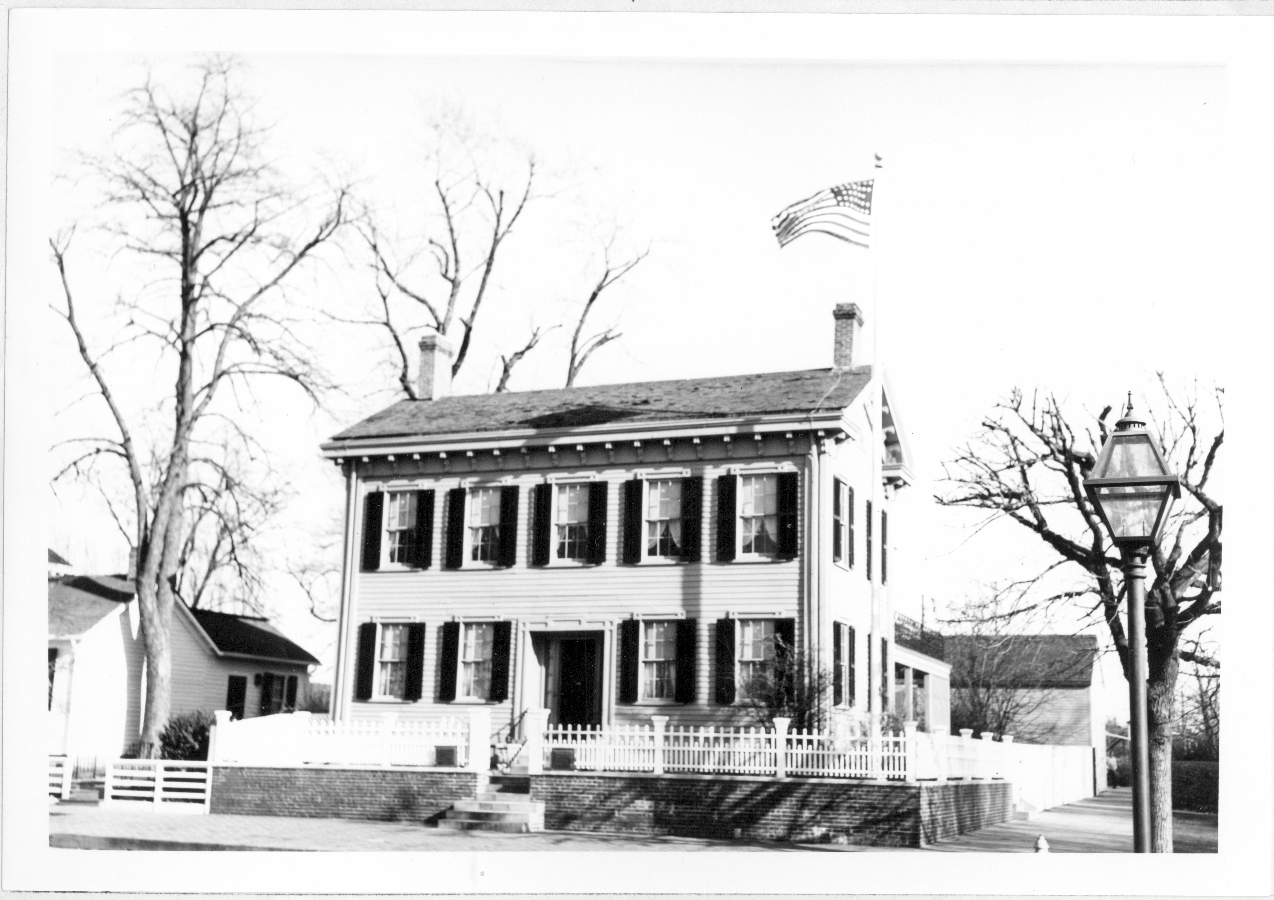 Lincoln Home west (front) elevation in winter, with flagpole in corner behind picket fence in corner. Lincoln Barn visible behind Home on right, Corneau house on left. Square commemorative plaques on either side of open gate. House appears to be painted white. Street light in foreground right. Looking East/Northeast from west side of 8th Street. Lincoln Home, Corneau, Lincoln Barn, painted white, 8th Street