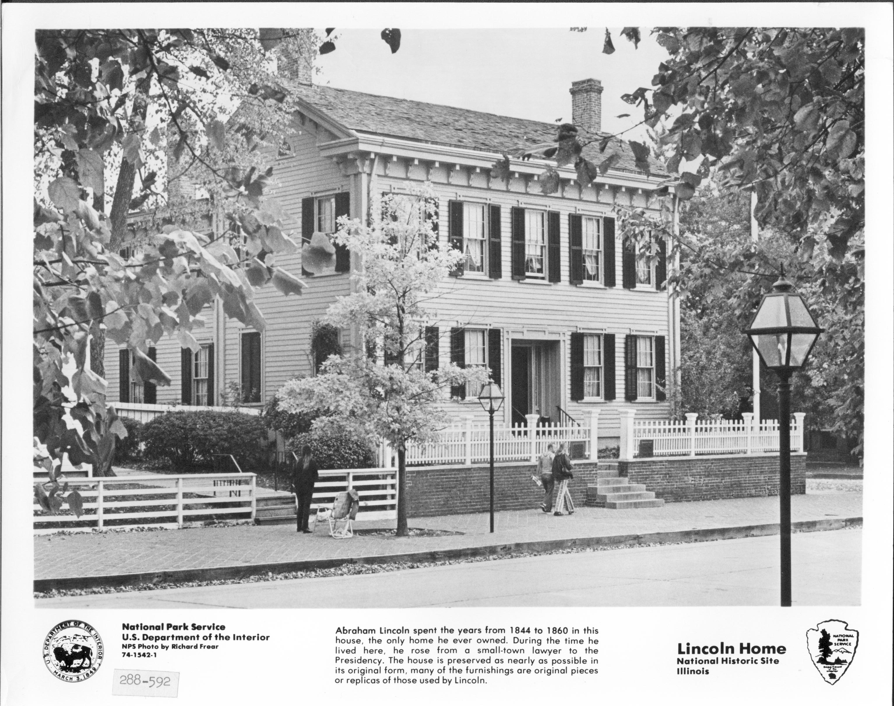 Lincoln Home west (front) elevation in official press photo. Features visitors walking past house and other visitor near a lawn chair on left. Flagpole behind picket fence in corner. Square commemorative plaques on either side of gate. DOI seal and NPS arrowhead are printed below photo on either side.  Additional information IDs photo as being from the NPS and DOI, and includes a brief description of site. Additional ID label: 288-592 Looking Southeast from west of 8th Street Lincol Home, NPS, official, visitors, flagpole, painted white?