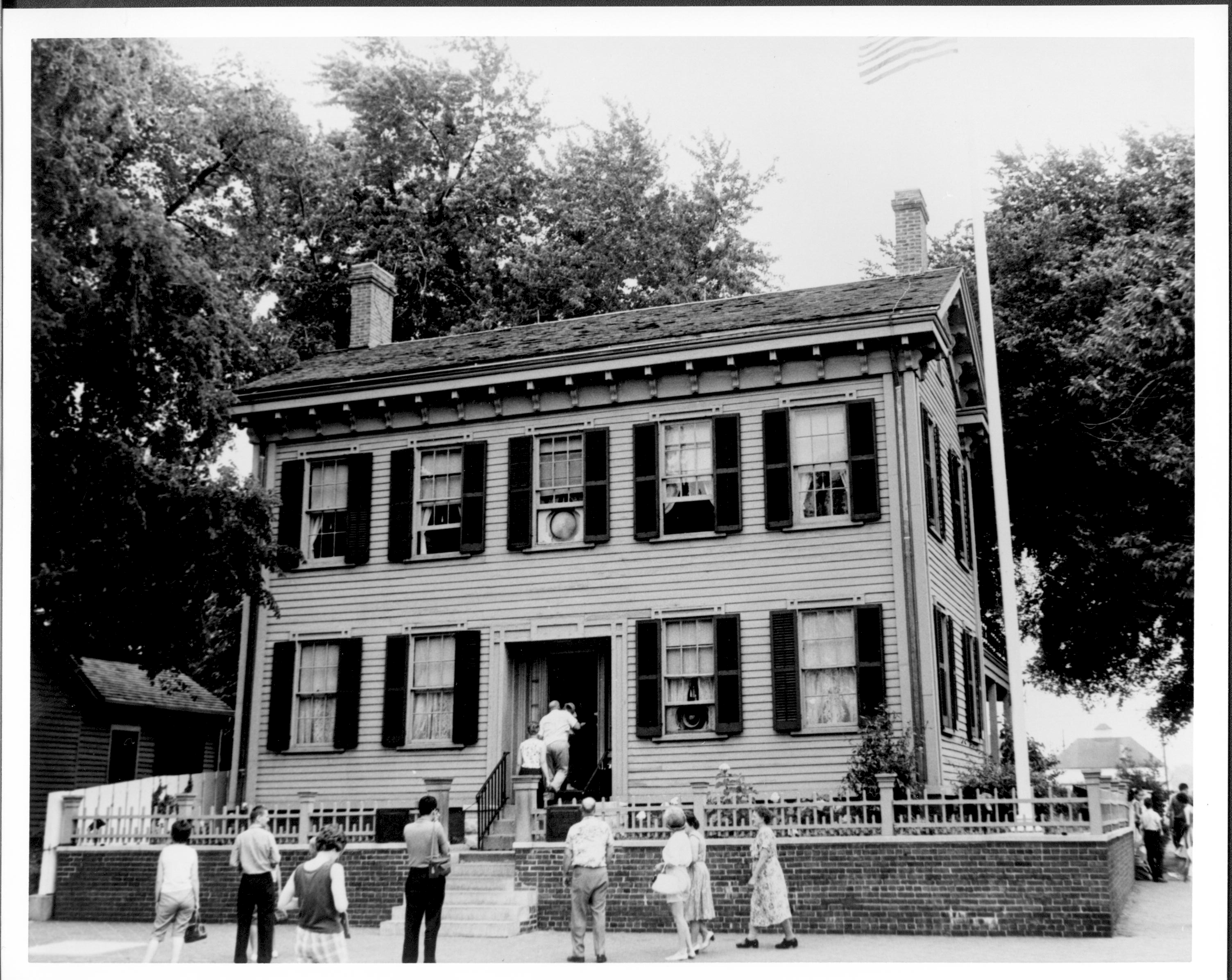 Lincoln Home front (West) elevation with visitors entering for a tour.  Windows are open and fans are visible in the upstairs center window and first floor right of the door.  A tall flagpole with a US flag flying is behind the fence on the corner.  Visitors are standing around in front of the Home and on the boardwalk on far right background. 3 people enter the house through the front door.  Corneau House is visible on far left. Lincoln barn visible in far background right. Looking East from 8th Street Lincoln Home, visitors, Corneau, 8th Street, Lincoln Barn, flagpole