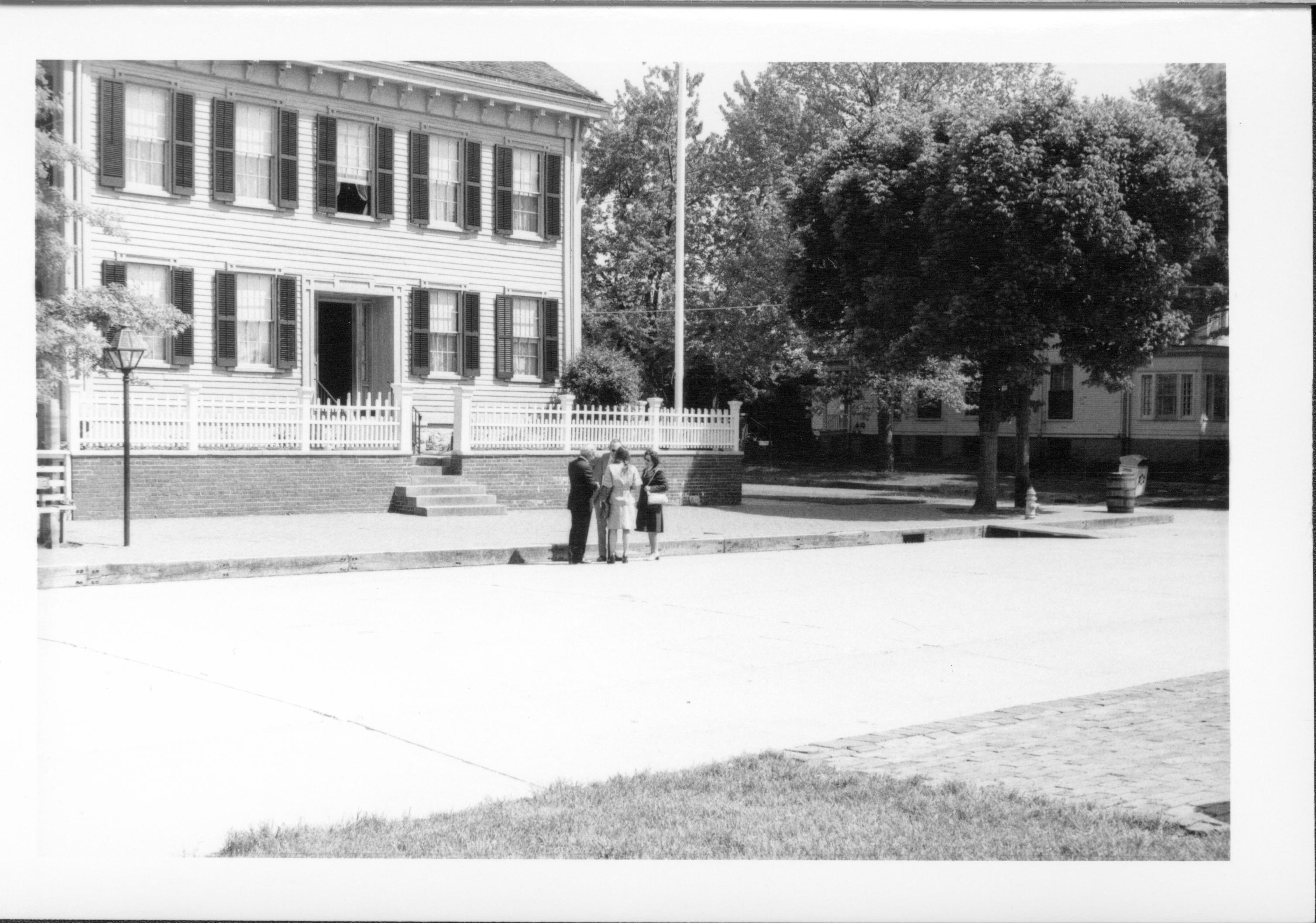 Lincoln Home front (West) elevation with small group of dressed-up visitors chatting in street in front of Home. Front door appears to be open, window above it is open.  Flagpole in corner behind fence is visible.  Rebecca Cook house visible in background right behind trash barrel and trash can on corners of 8th and Jackson Street intersection. Looking Southeast from west of 8th Street Lincoln Home, Rebecca Cook, 8th, Jackson, flagpole, visitors