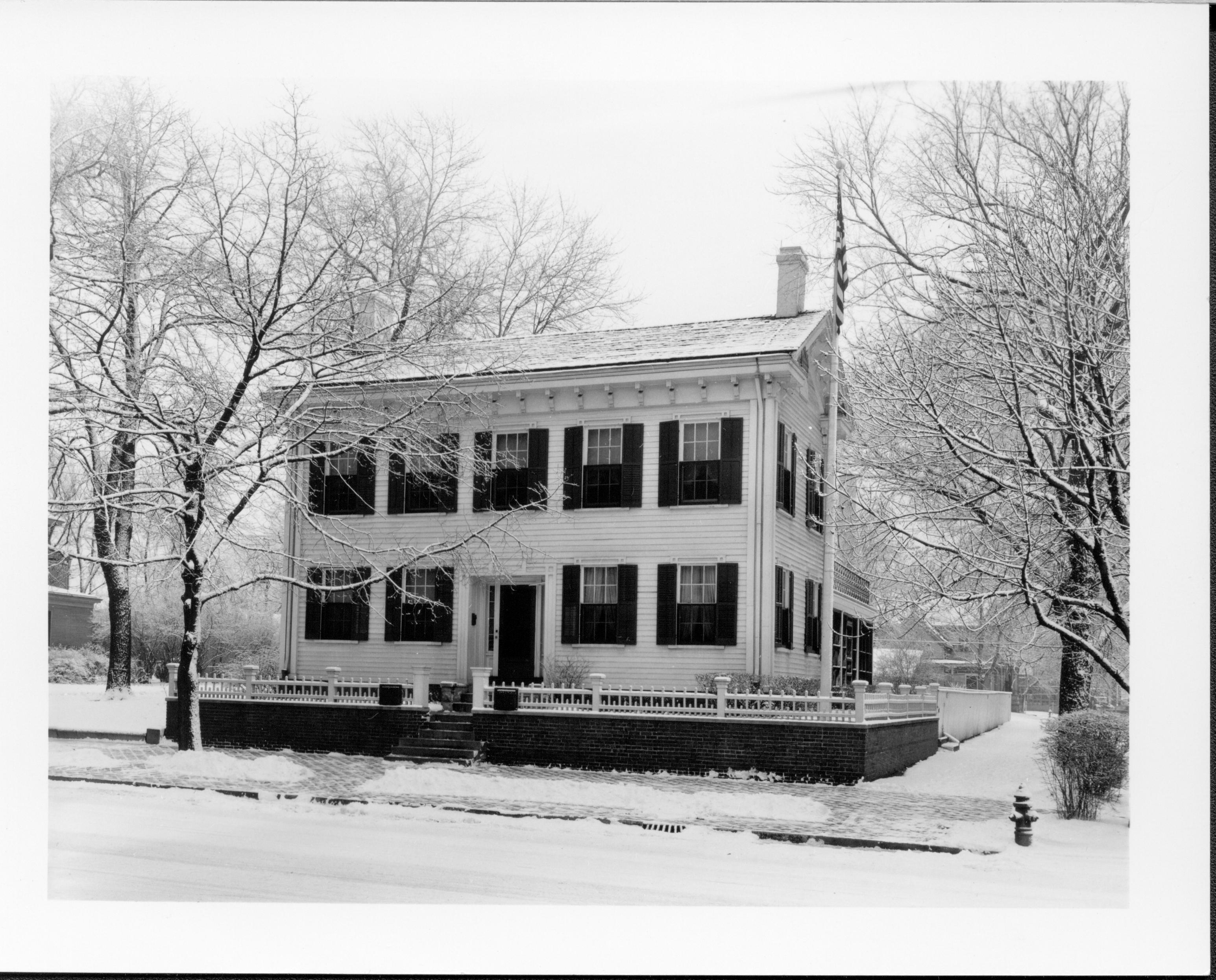 Lincoln Home west (front) elevation in winter, with flagpole in corner behind picket fence. Fire hydrant on corner of brick plaza.  Two story houses on Bugg lot (Block 10, lot 5) on far left, and behind in Kercheval lot (Block 10, lot 9) in background right. Back porch screened in. Looking East/Northeasat from 8th Street Lincoln Home, snow, Bugg, Kercheval, brick plaza, flagpole, screened back porch