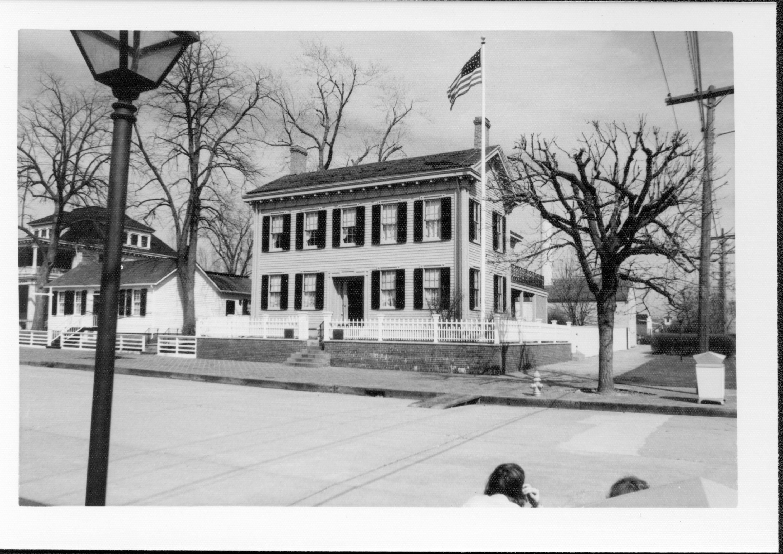 Lincoln Home front (West) elevation.  Visitors crouch down in front of trash can in foreground. Corneau House on left, large house on Bugg lot on far left.  Lincoln Barn in backgroudn right behind Home. Eletrical poles on far right along Jackson Street. Modern trash can on corner. Looking Northeast from 8th and Jackson Street intersection Lincoln Home, Corneau, Bugg, visitors, 8th, Jackson, electric pole, flagpole, Lincoln Barn